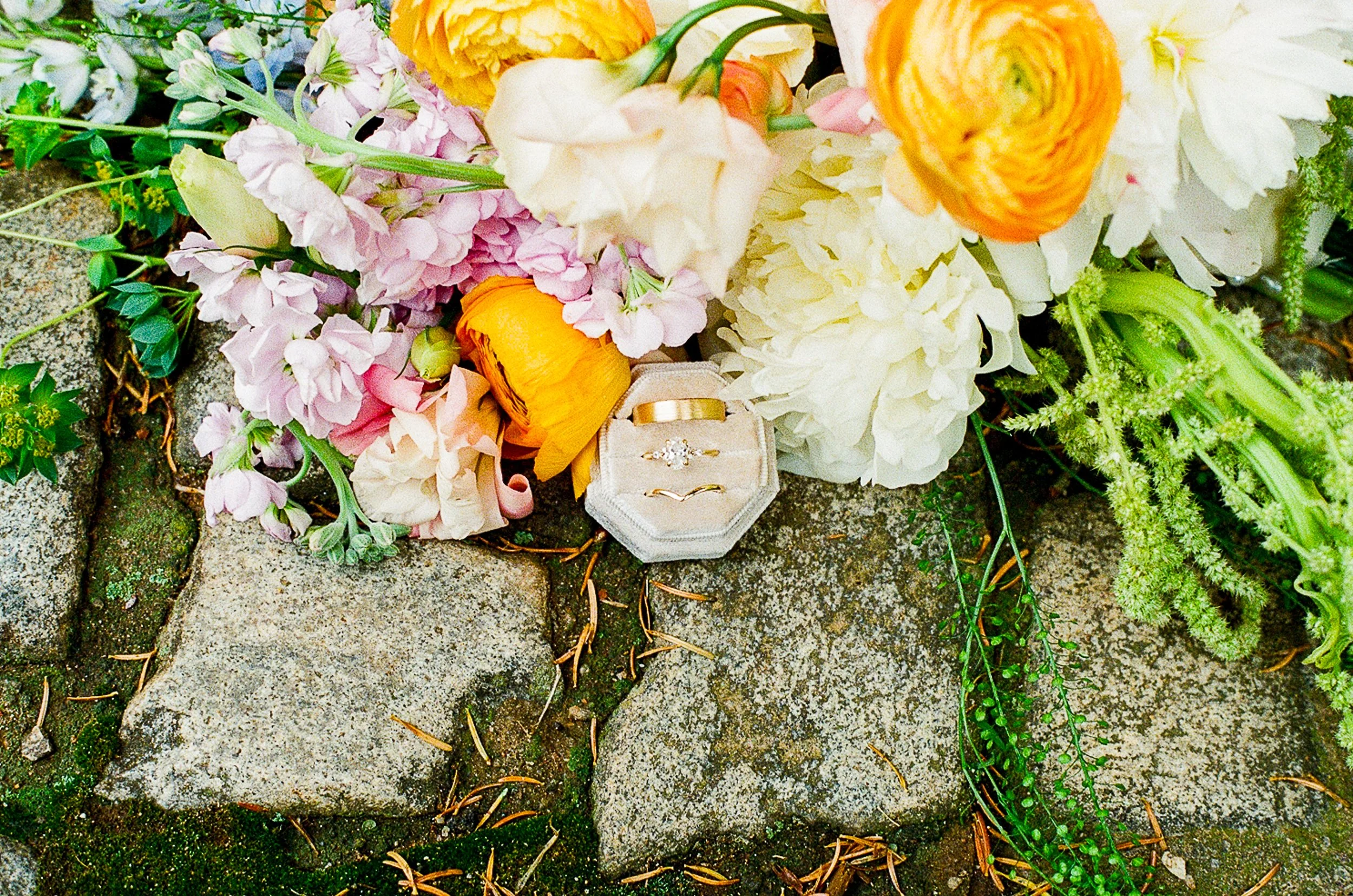 Close-up film photograph of colorful wedding florals arranged on a mossy stone path, featuring orange, pink, cream, and white blooms. A velvet ring box displaying three gold rings sits among the flowers.
