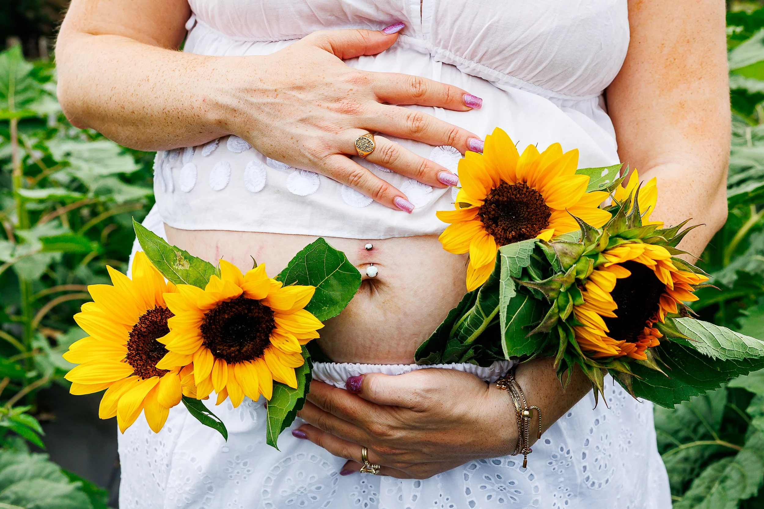 Close-up of a pregnant belly wrapped in a white dress, hand resting on top with two sunflowers held against one side, at Chiles Peach Orchard in Crozet, VA.