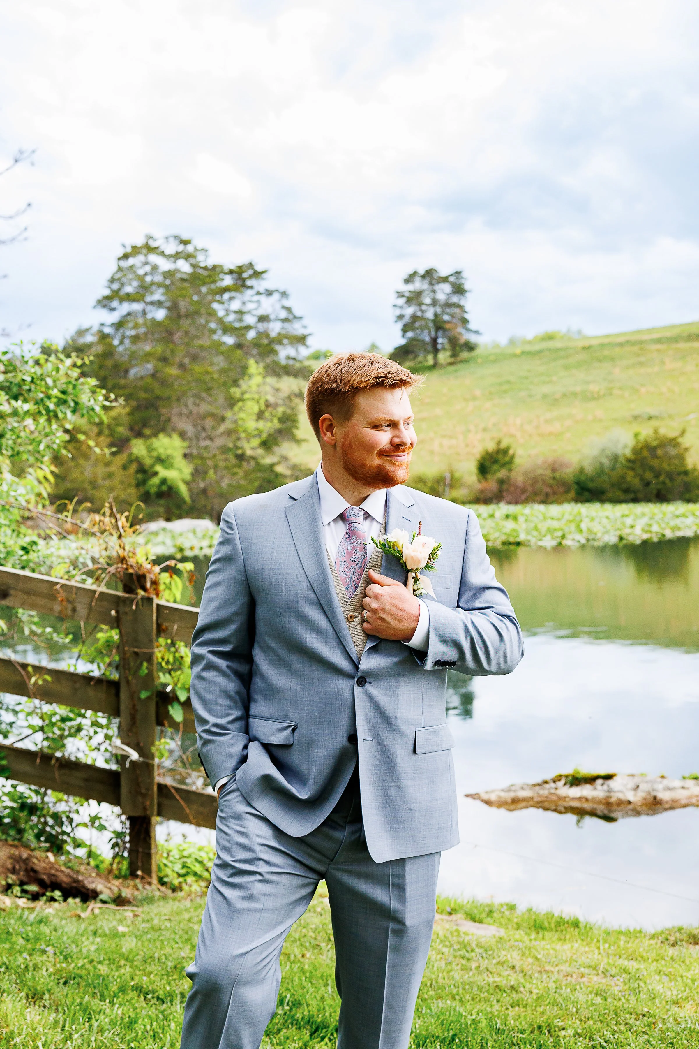 Groom in a light gray suit stands by a peaceful pond during an outdoor wedding, smiling off to the side with one hand in his pocket and the other adjusting his blush rose boutonniere. He wears a white shirt, pastel pink floral tie, and beige vest. A 