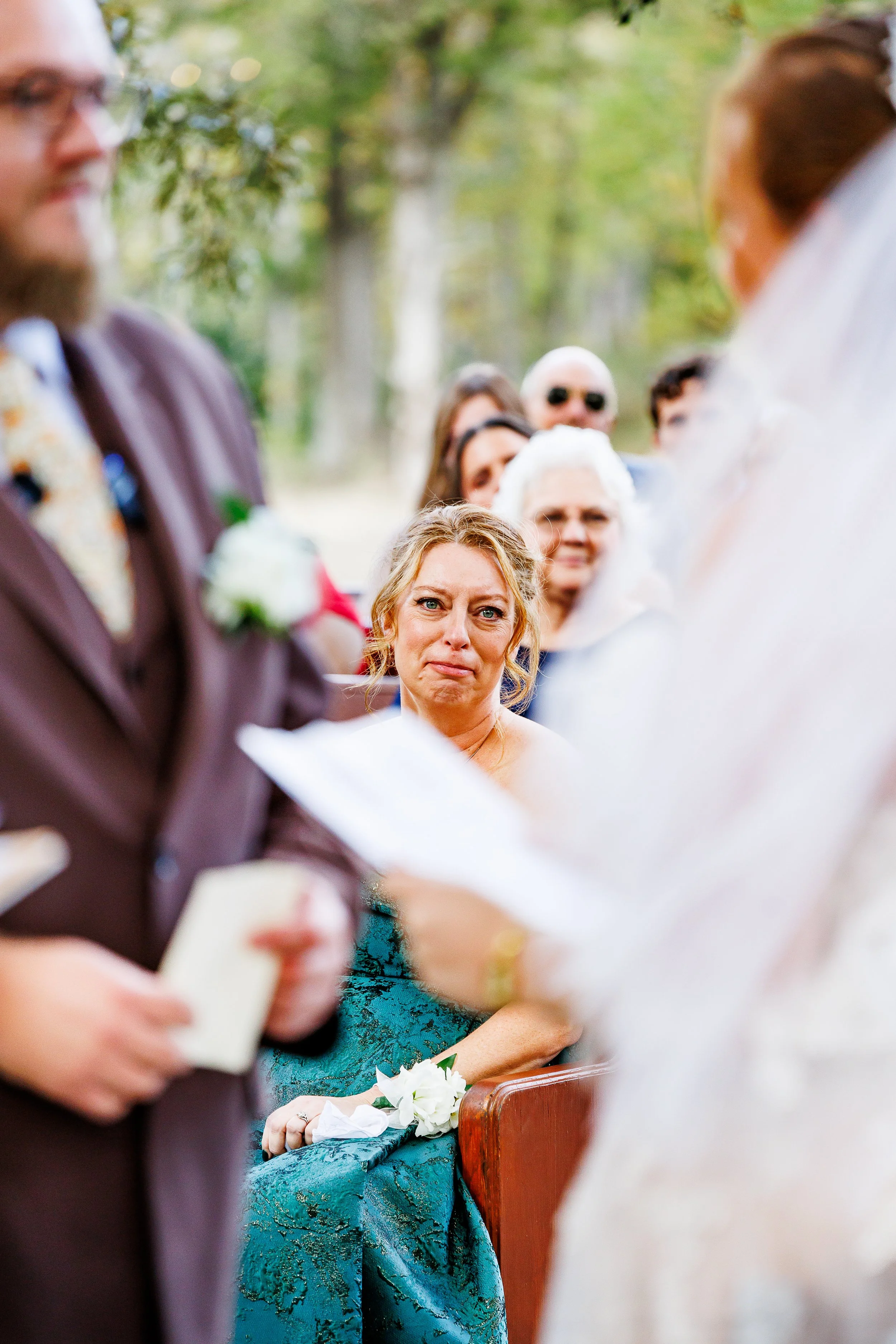 An emotional mother of the bride sits during an outdoor ceremony, looking tearful and deeply moved as she watches her daughter exchange vows. The bride and officiant are softly blurred in the foreground, while guests sit behind her among trees, creat
