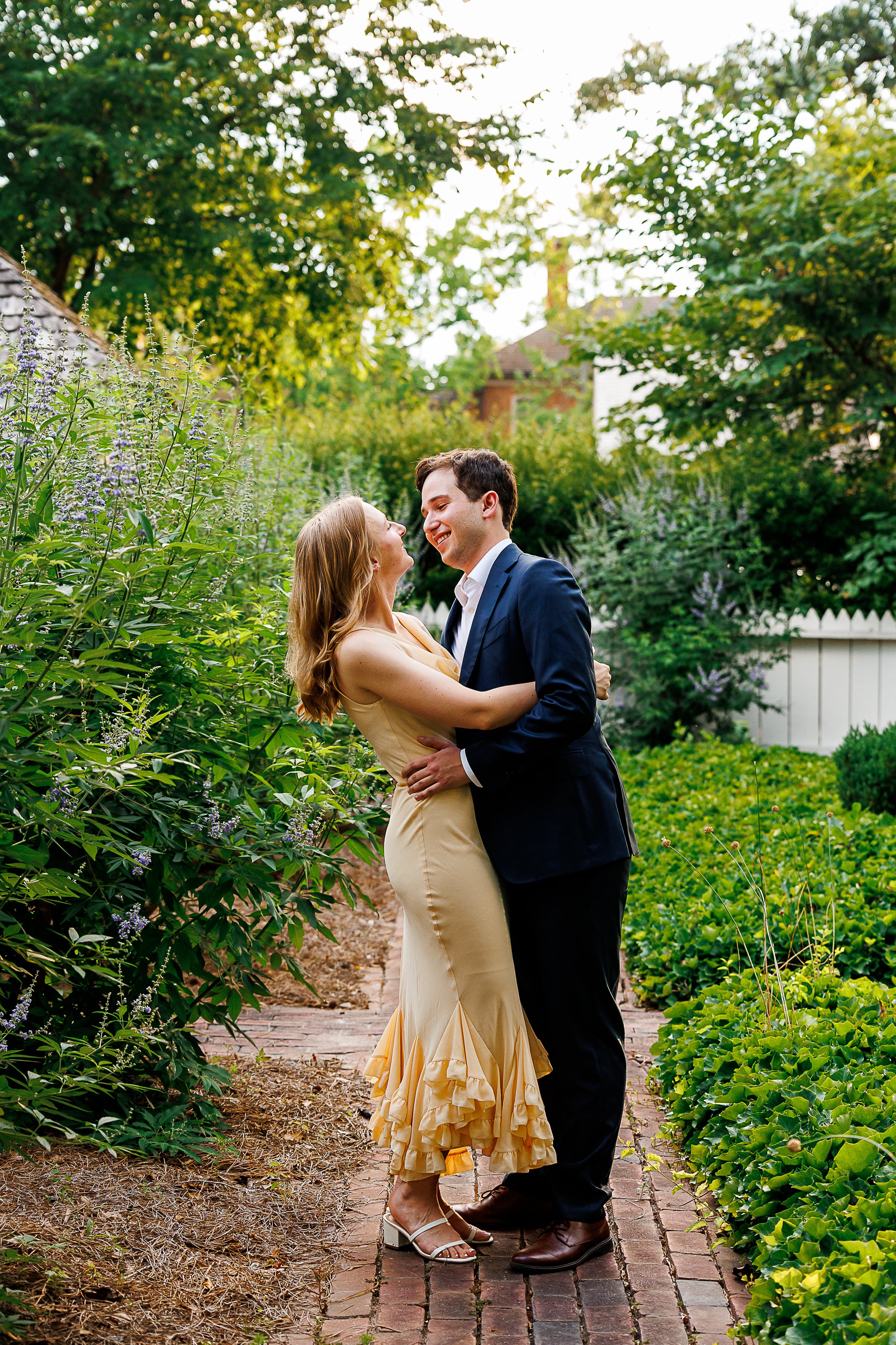Couple laughing and embracing on a brick garden path, her in a flowing yellow ruffled dress and him in a navy suit, surrounded by lush greenery and lavender blooms in Colonial Williamsburg, VA.