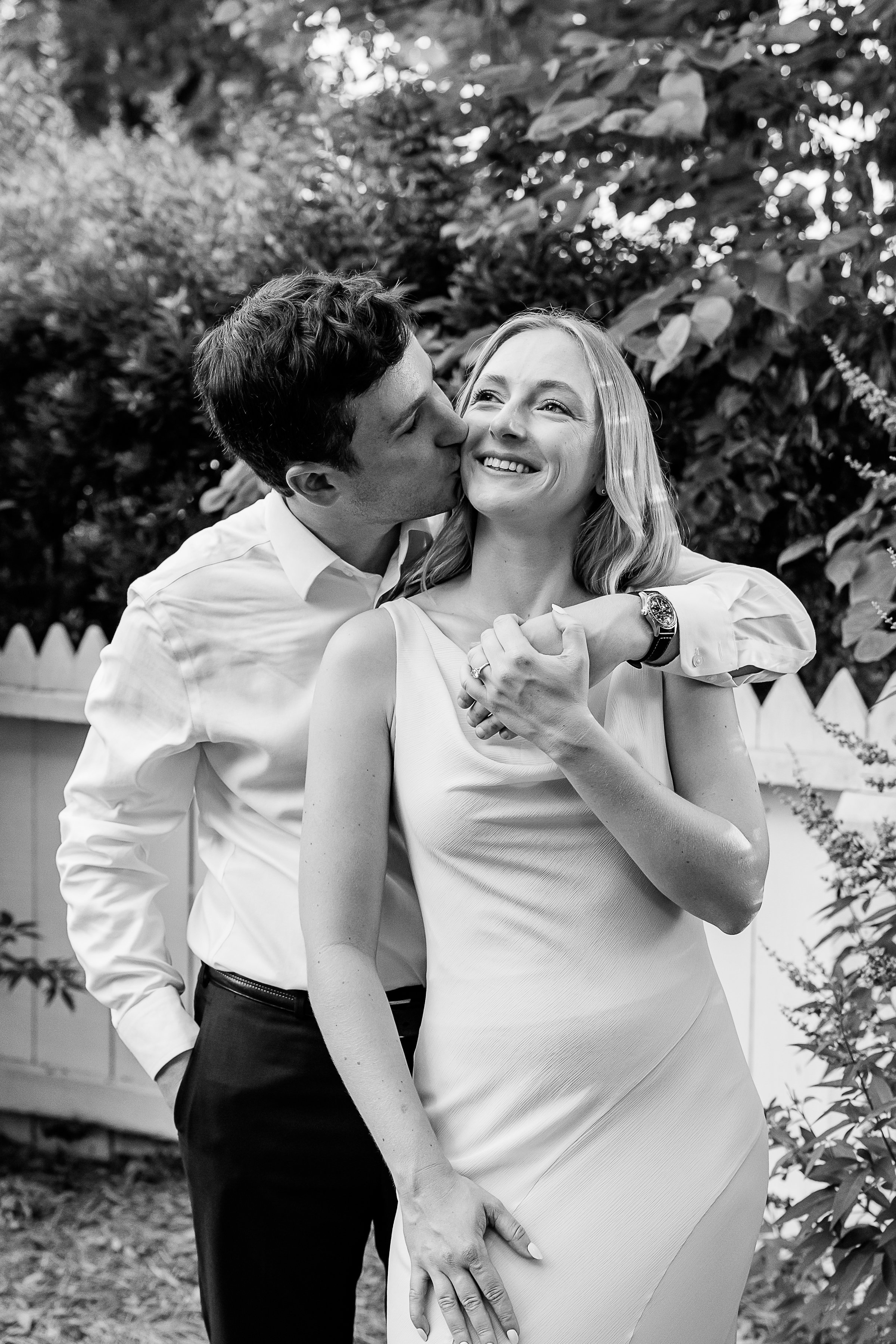 A black-and-white portrait of a couple sharing an affectionate moment in a lush garden. One partner stands behind the other with an arm wrapped around their shoulders, kissing their cheek while they smile brightly off into the distance. Their hands a