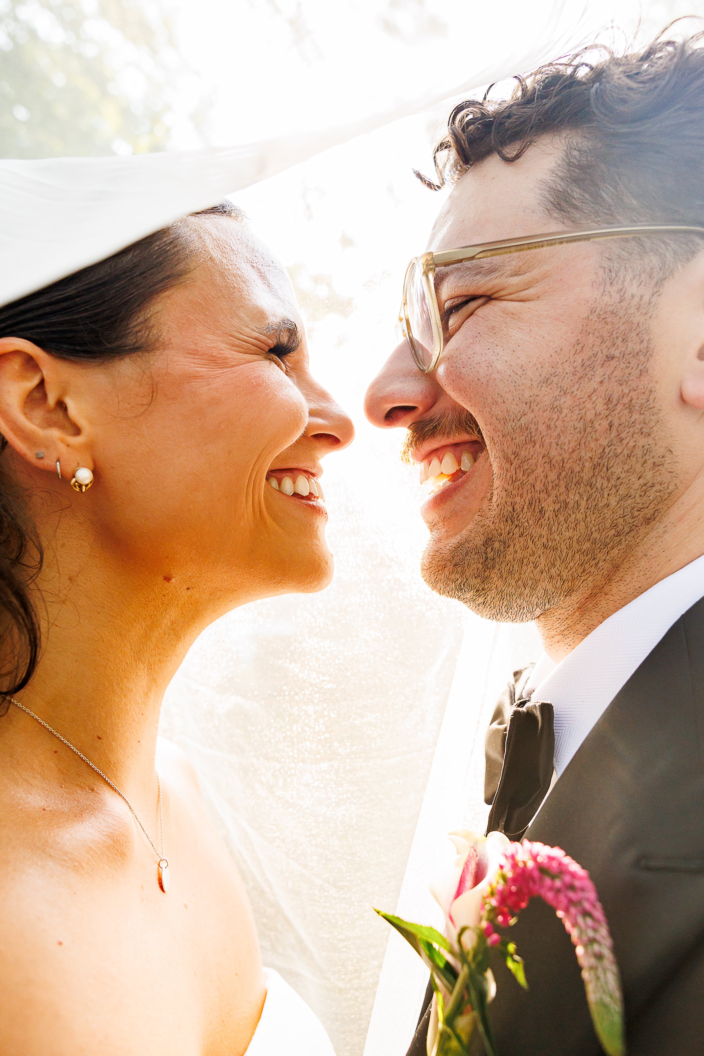 Bride and groom laugh nose-to-nose under the wedding veil in backlit sunlight at Virginia Cliffe Inn, groom wearing glasses with a pink floral boutonniere.