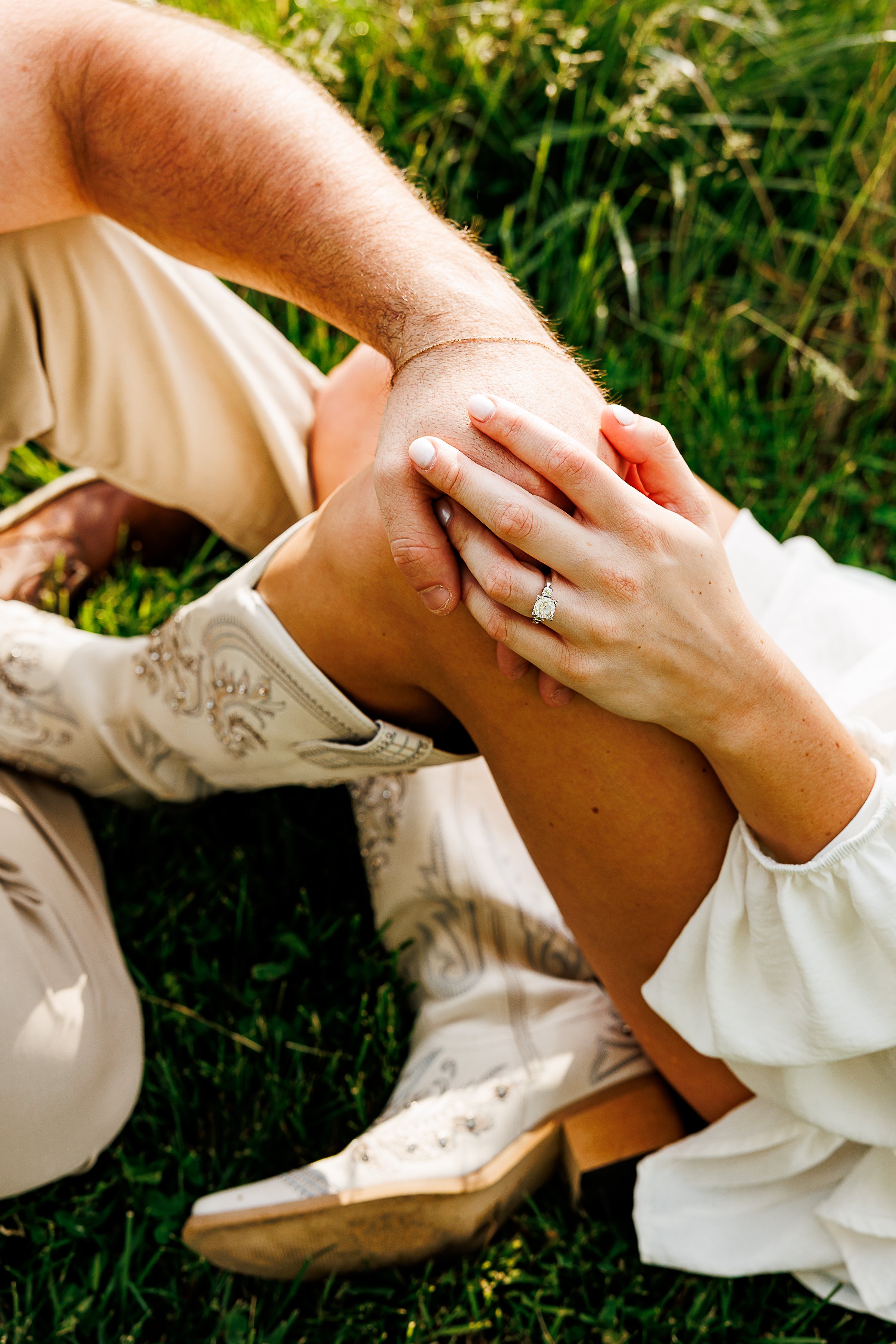 A close-up of a couple sitting together on the grass with their hands intertwined. An oval-cut engagement ring sparkles on one partner’s hand, while white embroidered cowboy boots and neutral-toned clothing add a western-inspired touch. Warm sunlight