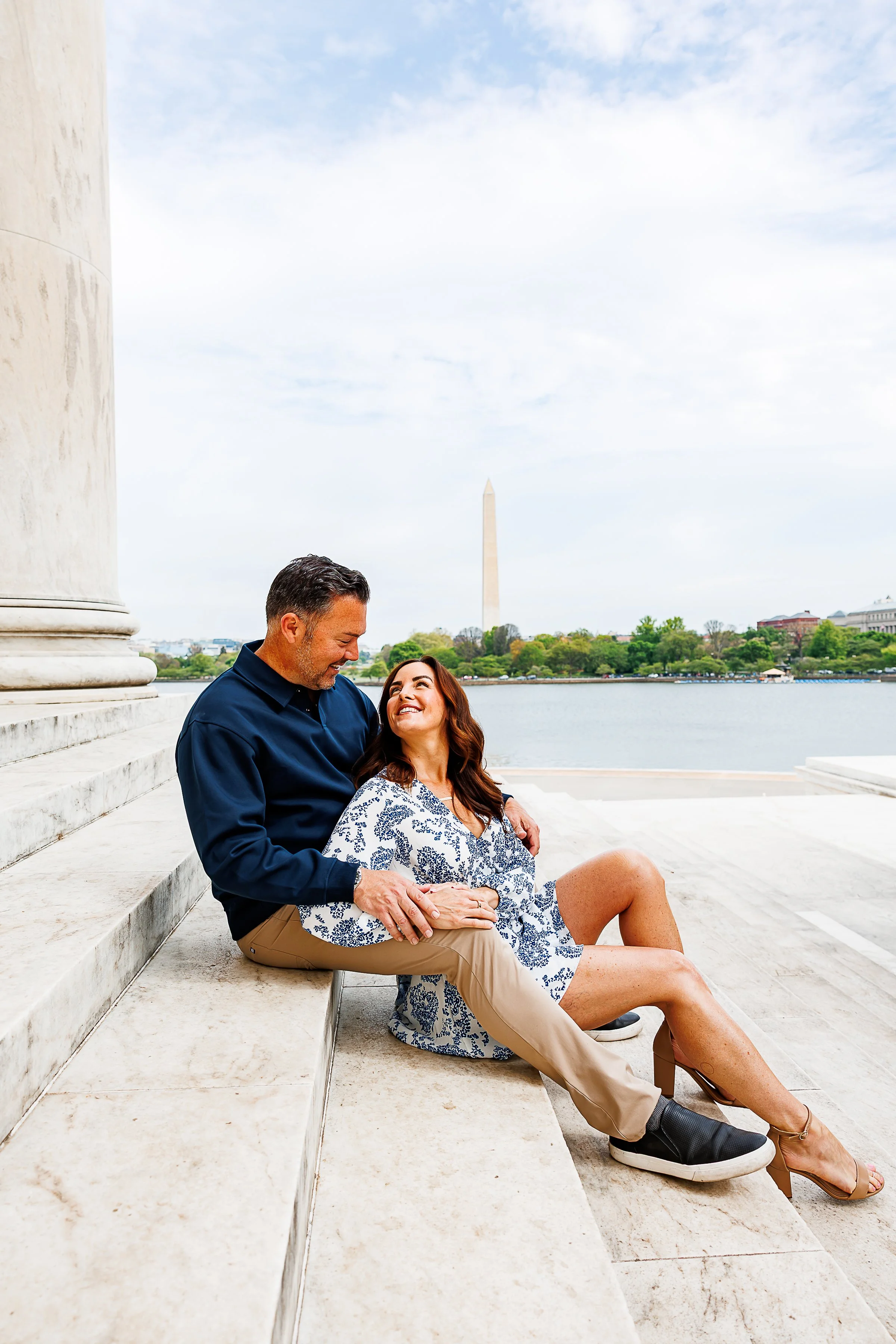 A couple sits together on the stone steps of a waterfront monument in Washington, D.C., with the Washington Monument rising in the background. The man leans in with his arm around the woman as she smiles up at him, both dressed in soft, neutral tones