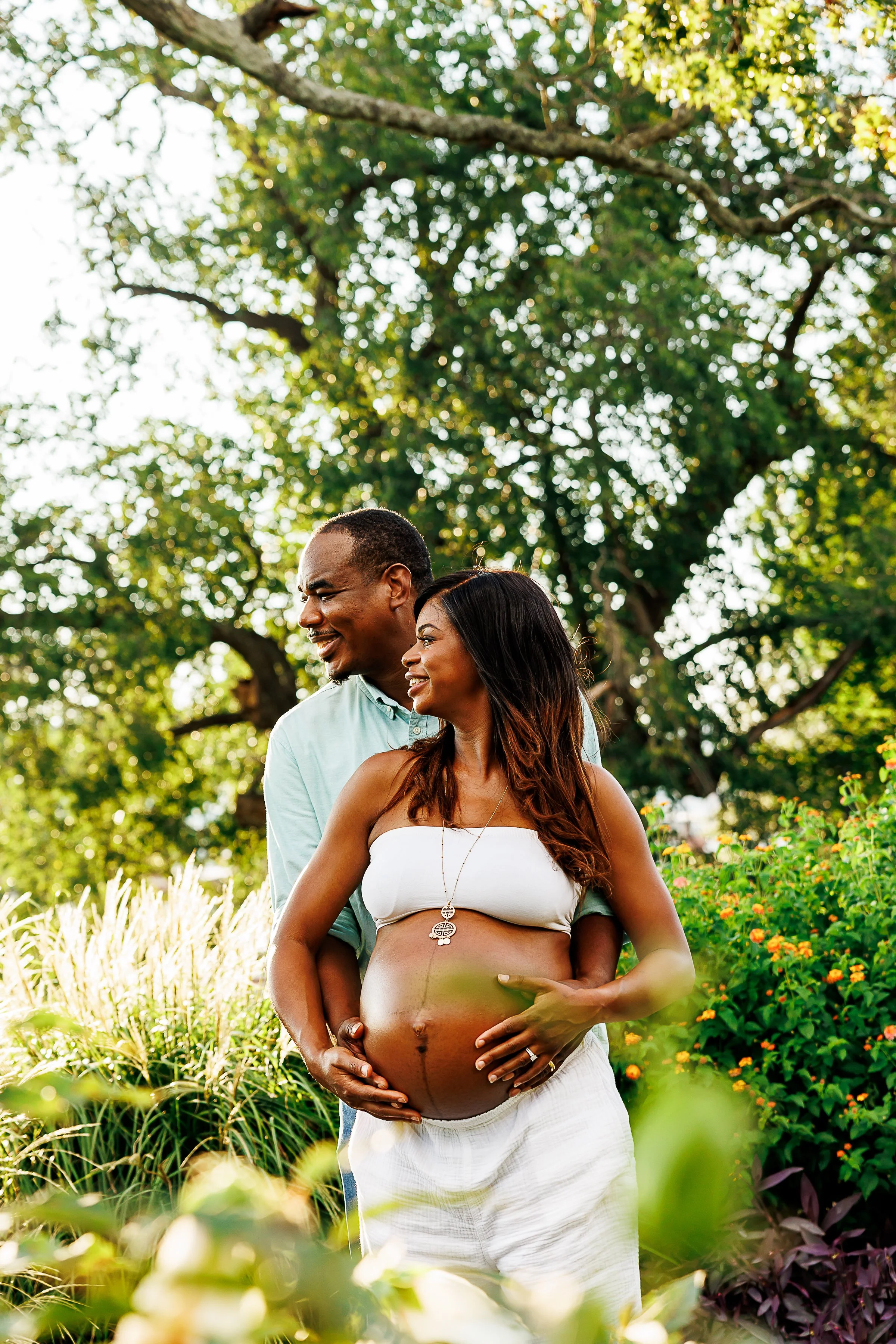 Pregnant woman in a white bandeau top smiling with her partner standing behind her, hands cradling her belly, surrounded by tall grasses and orange flowers at Libby Hill Park in Richmond, VA.