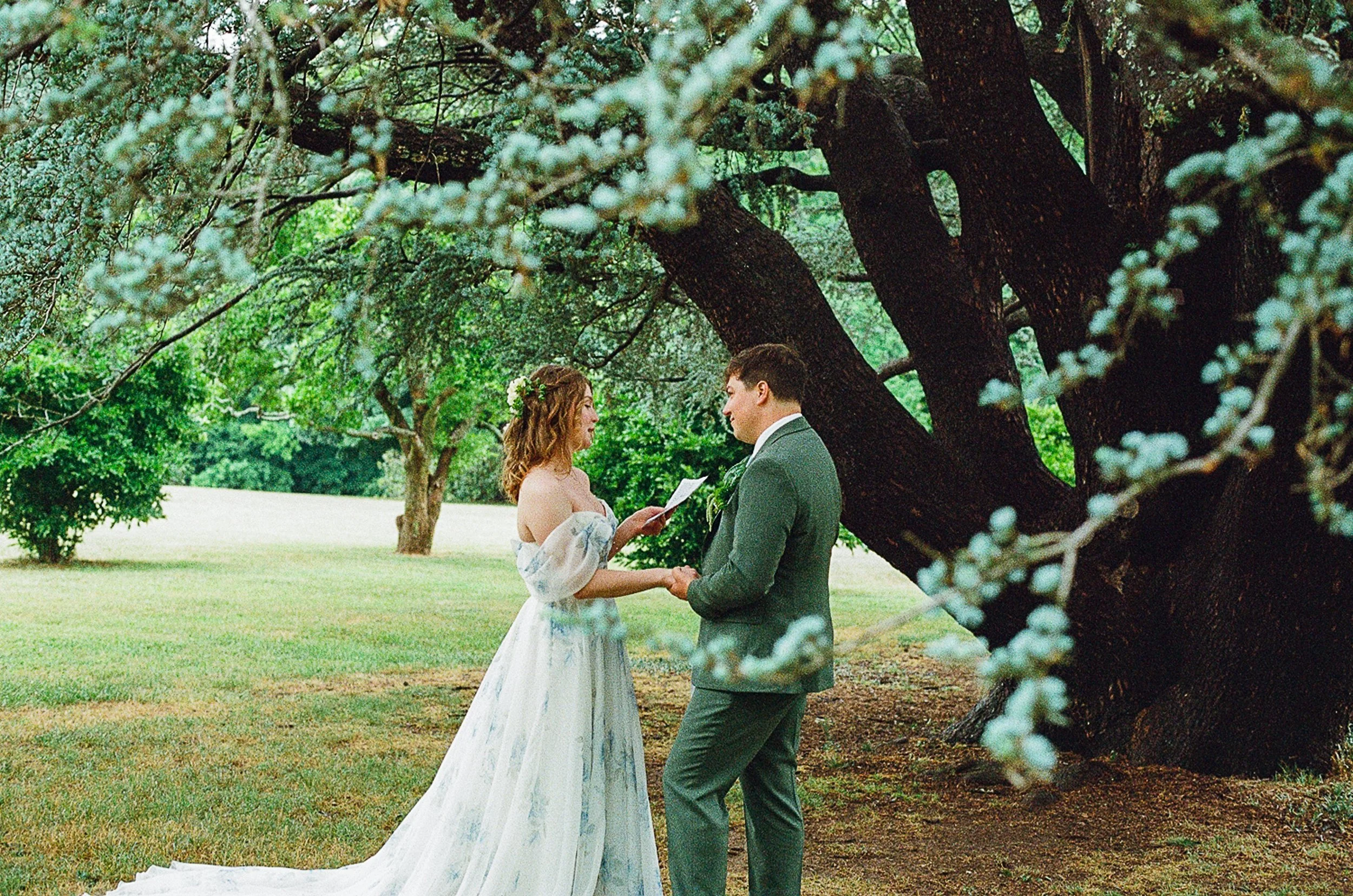 Film photograph of a bride and groom holding hands and exchanging private vows beneath a large tree. Soft greenery and blurred branches frame the couple in an intimate outdoor setting.