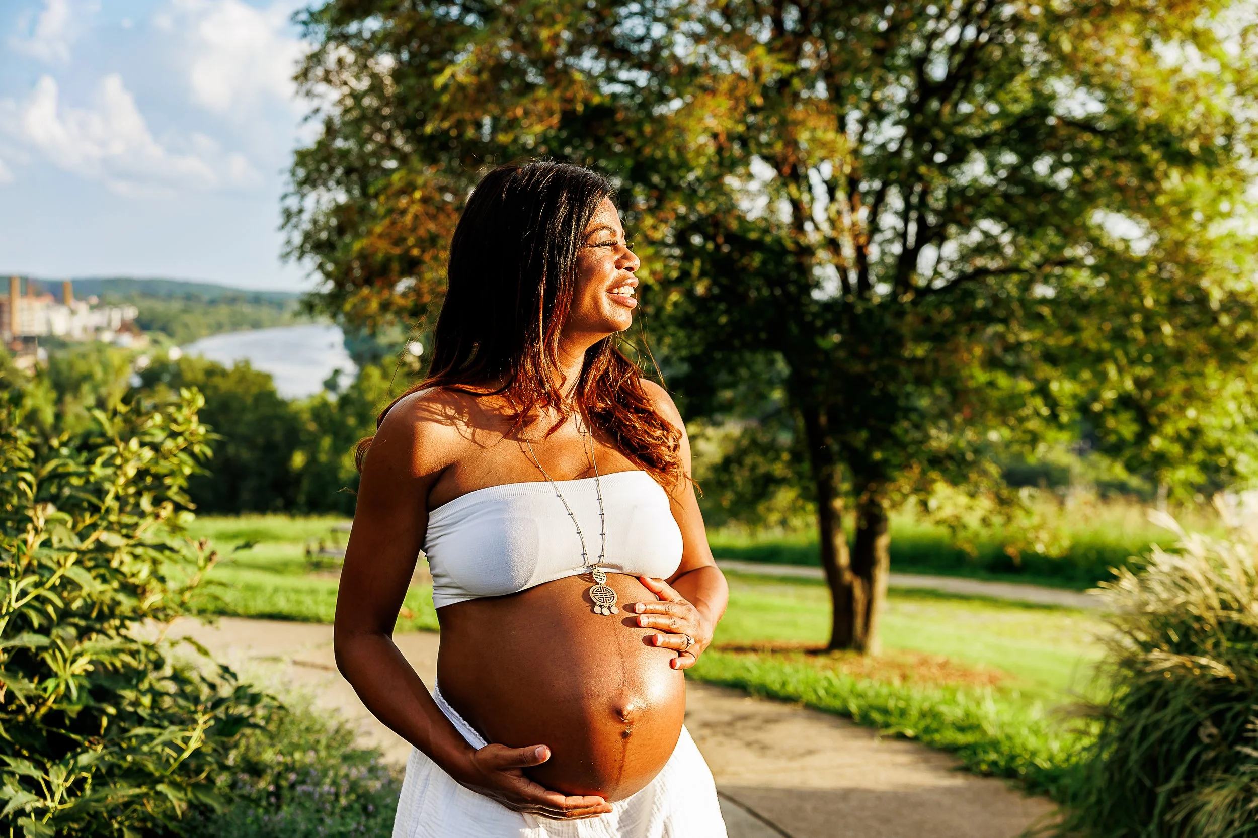 A pregnant woman stands outdoors in warm golden sunlight, gently cradling her baby bump with both hands. She wears a white strapless top and flowing white bottoms, with lush trees, greenery, and a river visible in the distance.