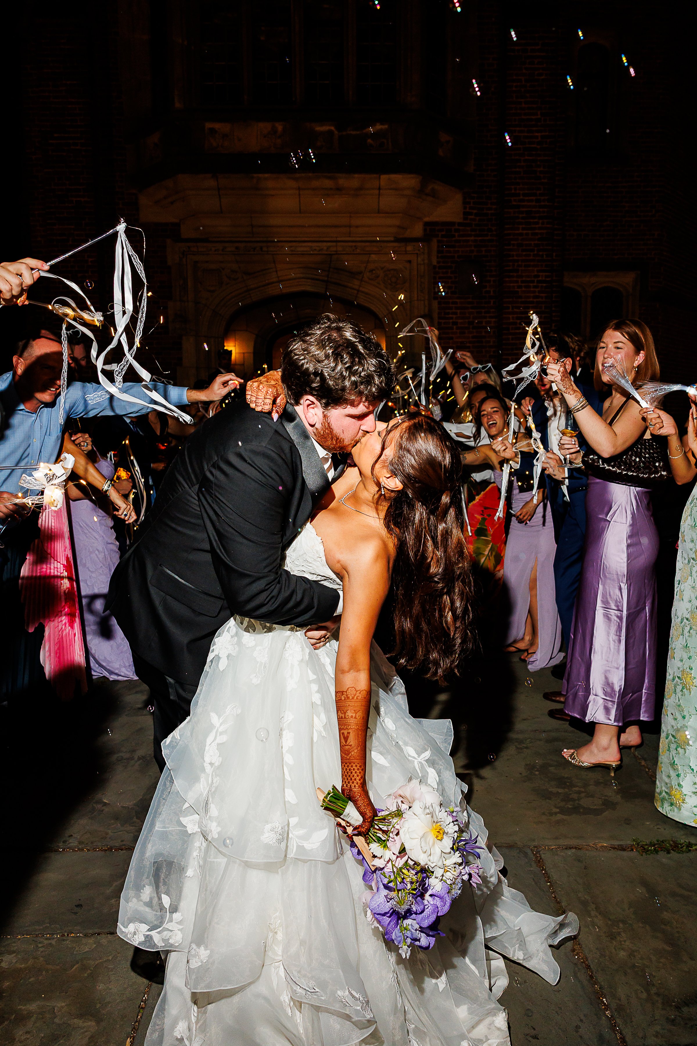 Bride and groom share a dramatic dip kiss during their nighttime exit as guests surround them waving ribbon wands and blowing bubbles. The bride holds a bouquet of white and purple flowers while her flowing gown and intricate henna designs stand out 