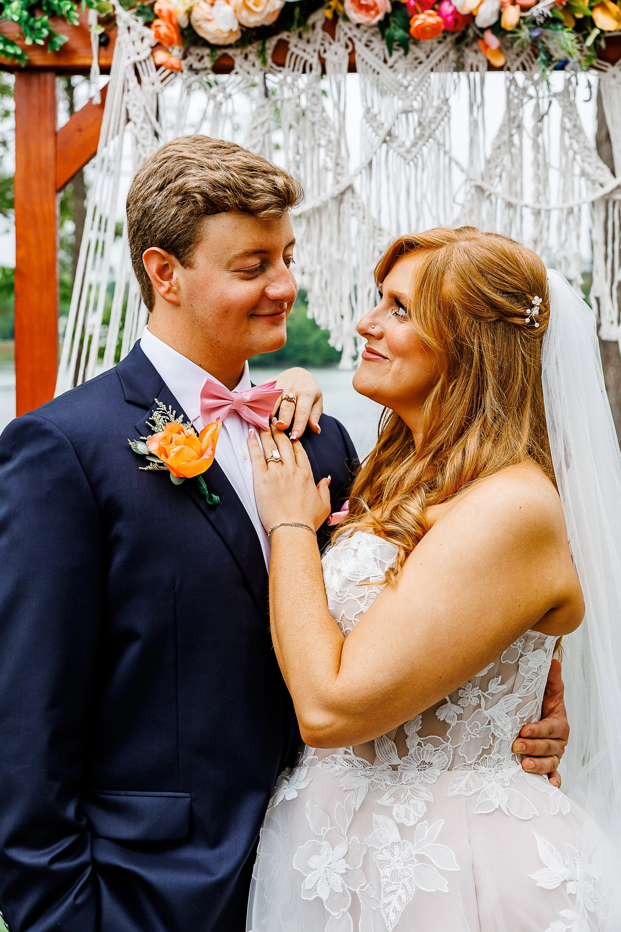 A newlywed couple shares a tender moment beneath a floral wedding arbor decorated with colorful blooms and macramé details. The groom wears a navy blue suit with a pink bow tie and a bright orange boutonniere, while the bride wears a lace wedding gow