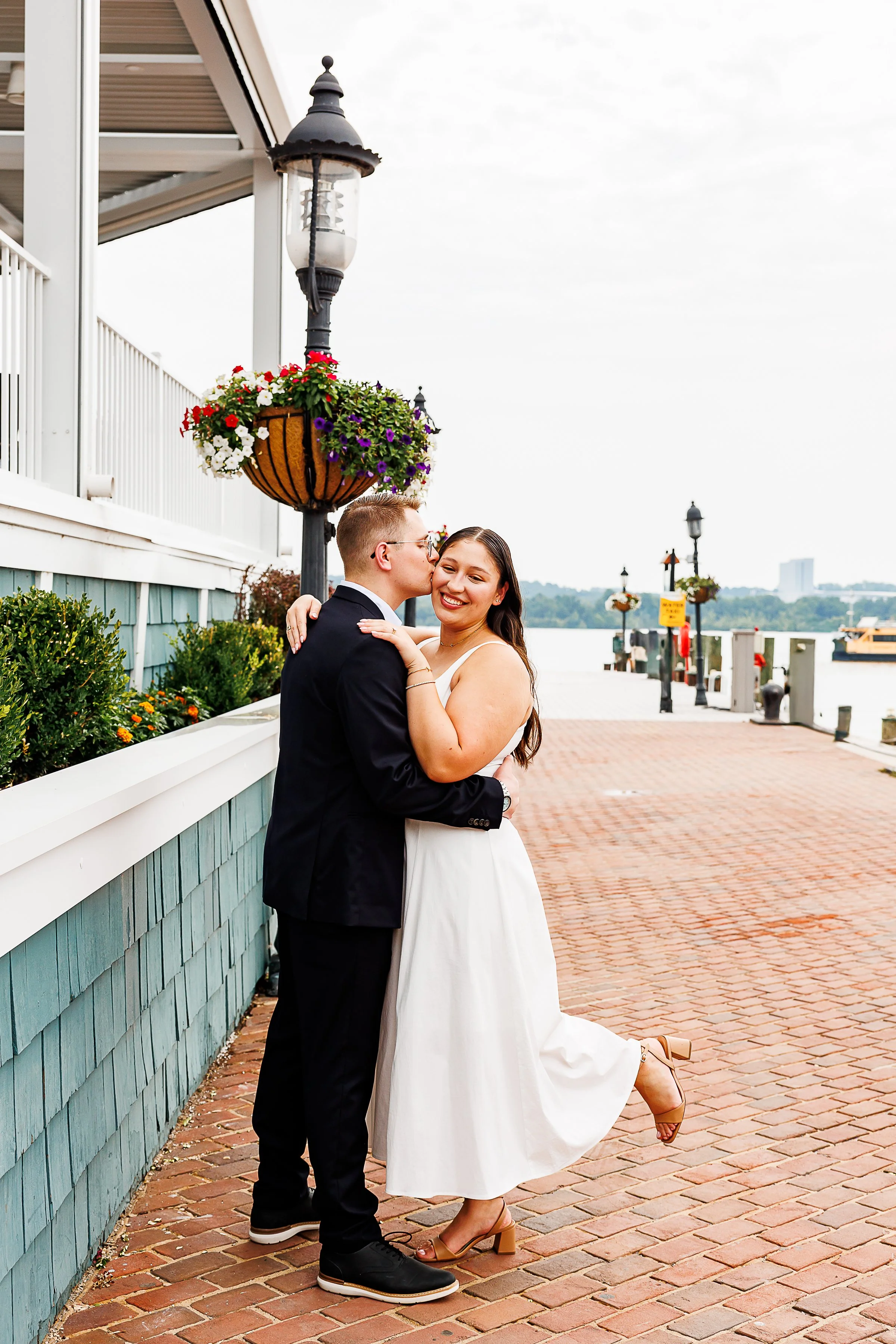 A couple embraces along a brick waterfront promenade as one partner kisses the other on the cheek. The other smiles toward the camera with one leg lifted playfully behind them. Hanging flower baskets, waterfront views, and classic lampposts create a 