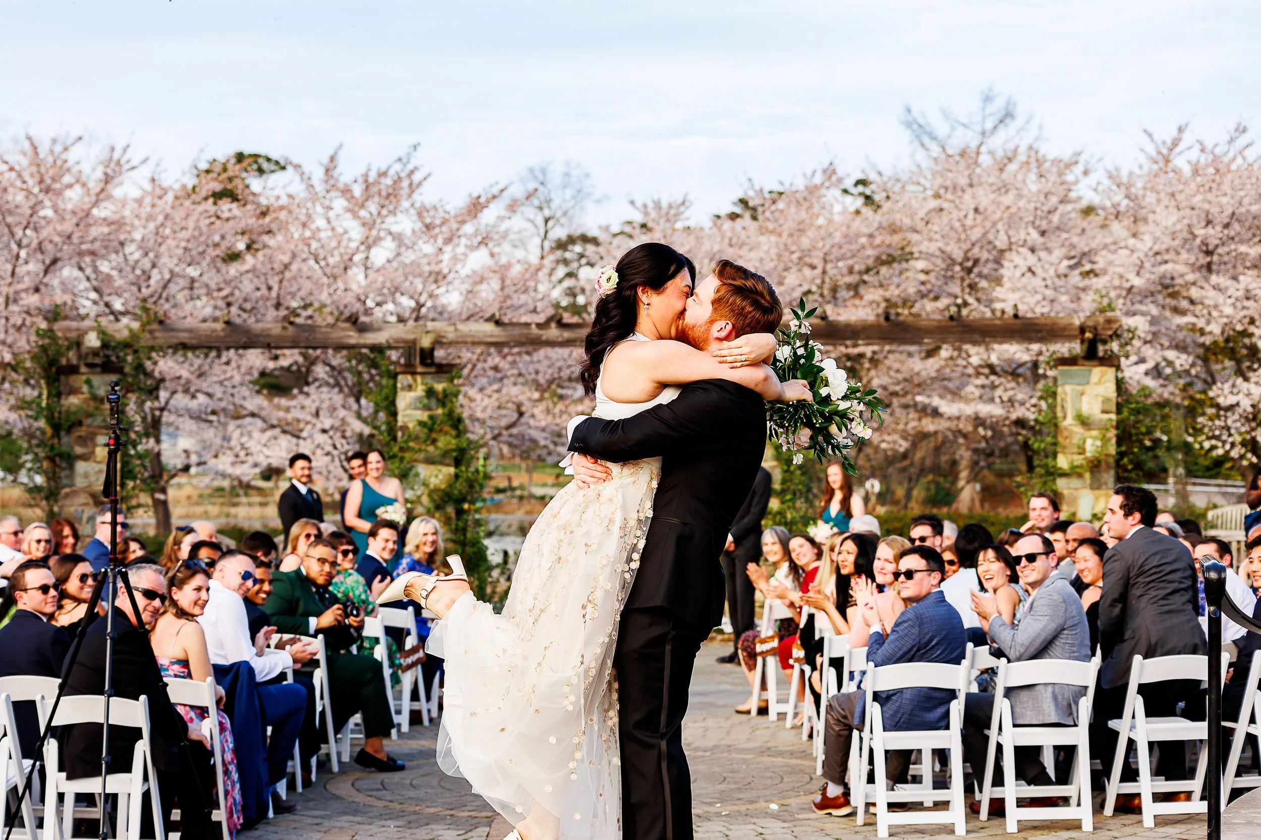 A joyful just-married moment as the groom lifts the bride into a kiss in the center of the aisle, surrounded by cheering guests. Rows of white chairs frame the scene while friends and family smile and applaud, and blooming cherry blossoms create a ro