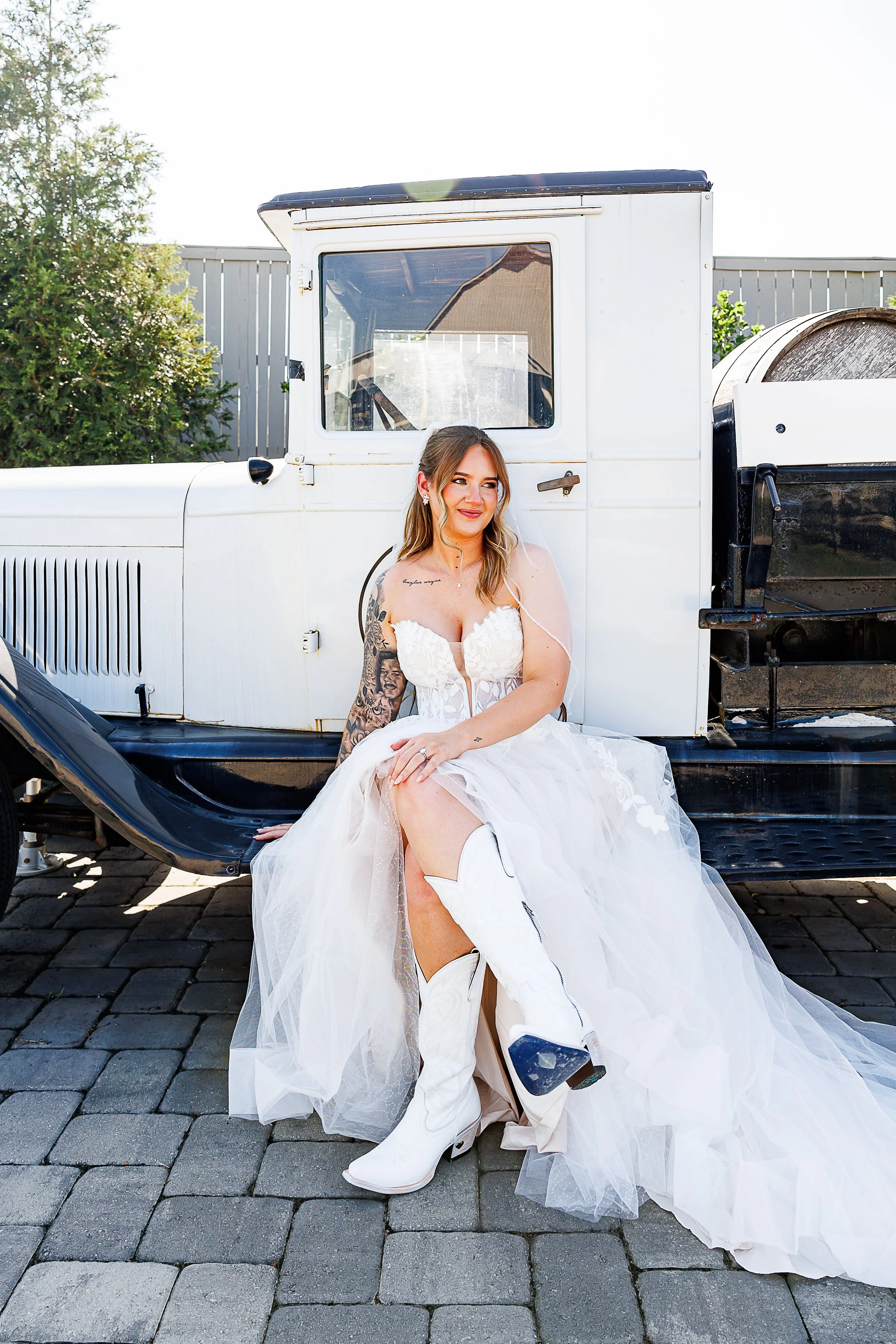 Bride in a strapless lace wedding gown and white cowboy boots sits on the running board of a vintage white truck, smiling off to the side. Her veil drapes over one shoulder as the tulle of her dress cascades onto the stone pavers below, creating a pl