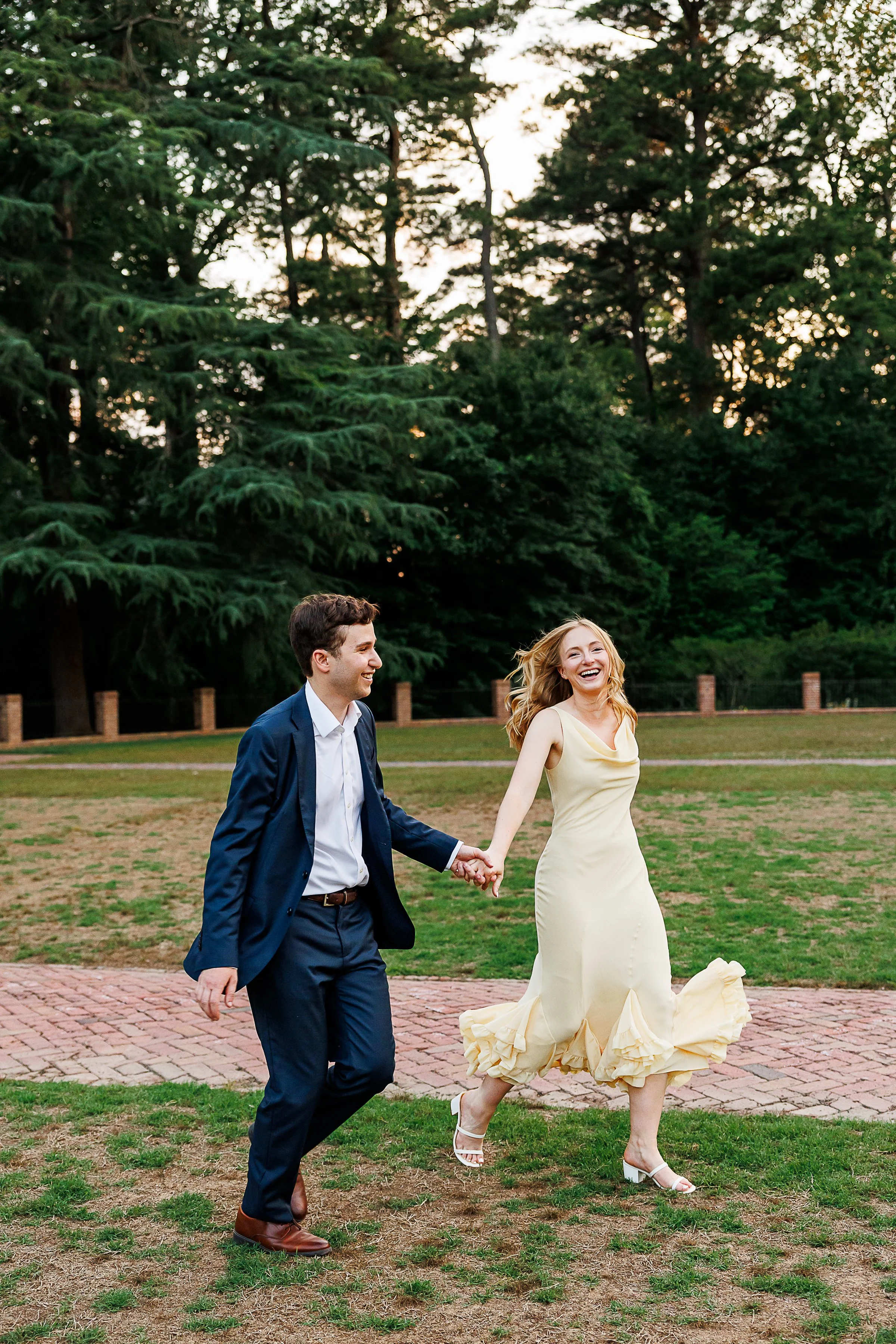 A smiling man and woman walk hand in hand outdoors on a brick path, with tall green trees and a sunset sky in the background. The woman wears a yellow dress and white heels, and the man wears a suit and brown shoes.