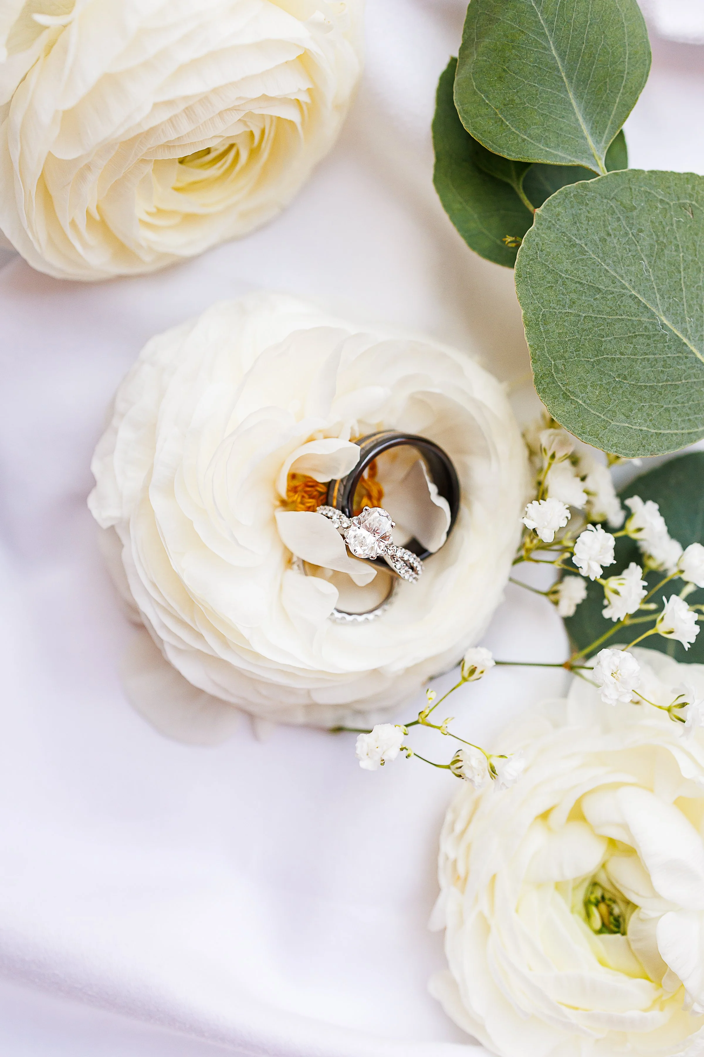 Close-up of white ranunculus flowers with green leaves, a pair of rings including a diamond engagement ring and a wedding band placed on one flower.