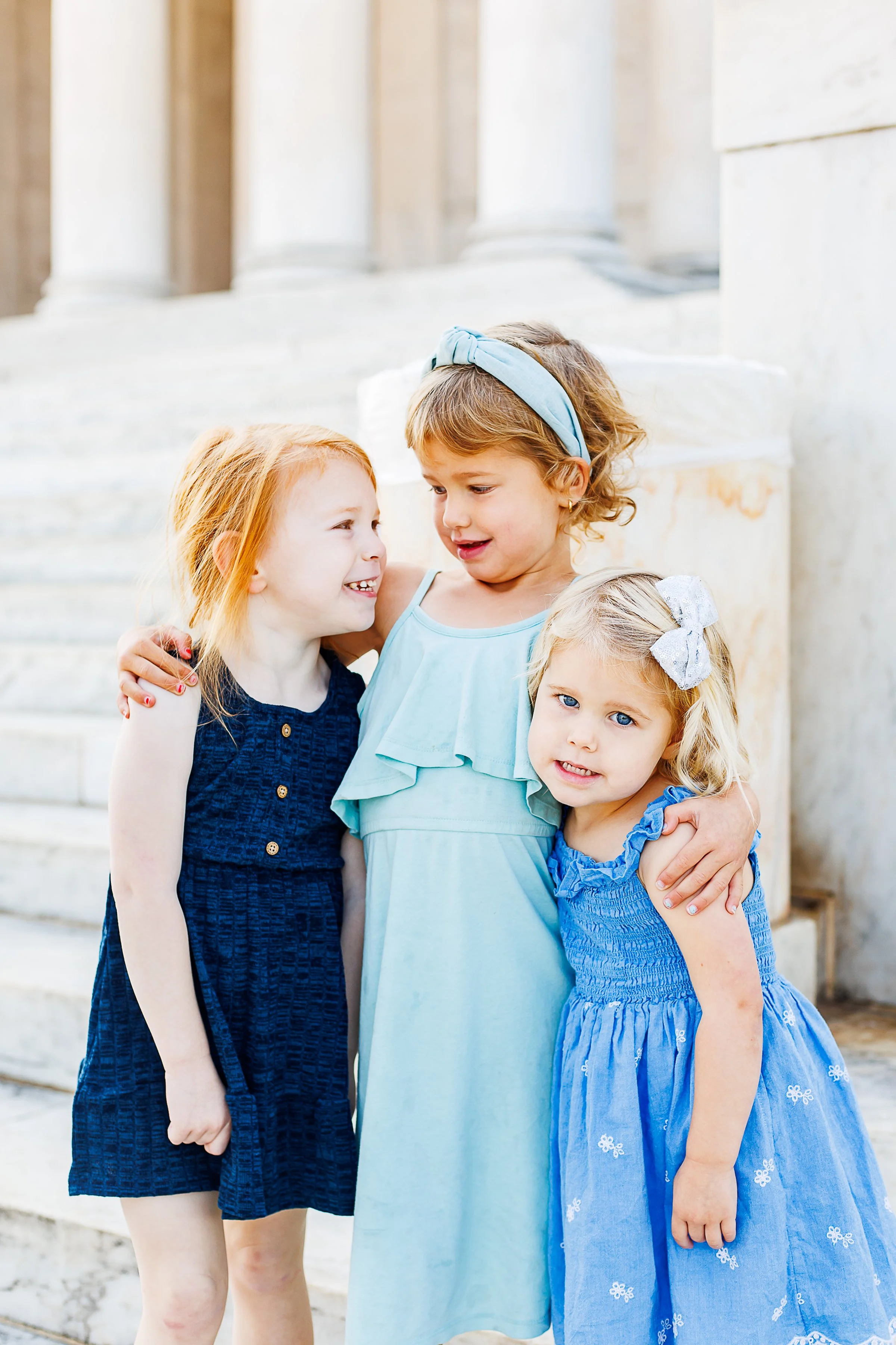 Three sisters in shades of blue dresses laughing together with arms around each other's shoulders, in front of the marble columns at the Thomas Jefferson Memorial in Washington, DC.
