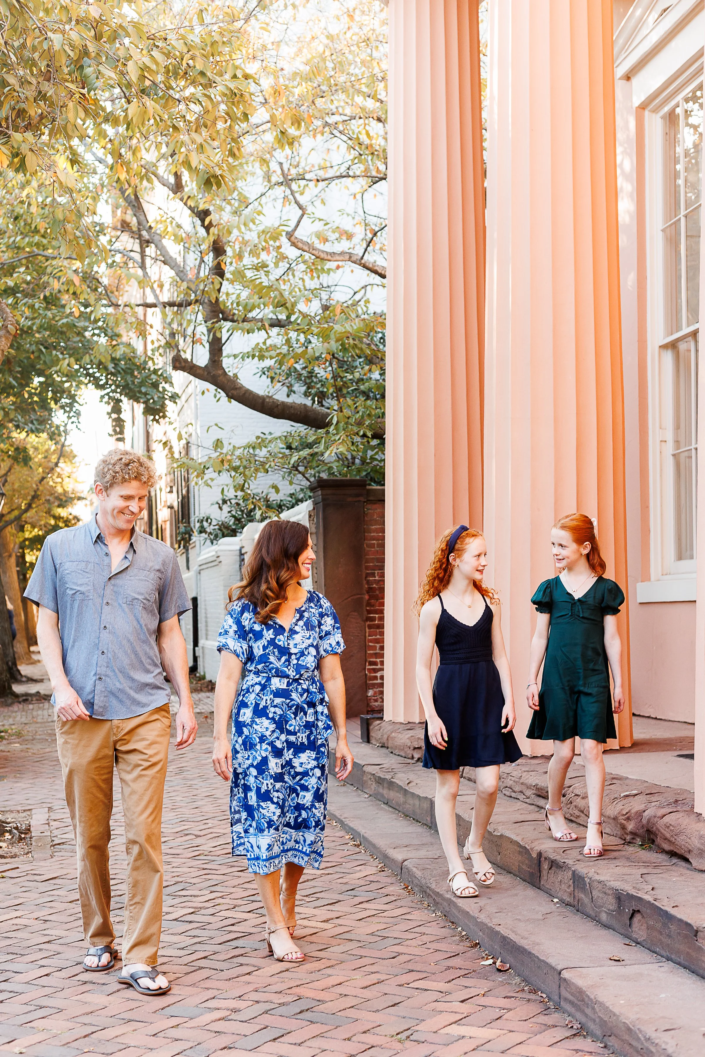 Family of four walking and laughing together down a brick sidewalk past a peach-colored colonial building with tall columns, in Old Town Alexandria, VA.
