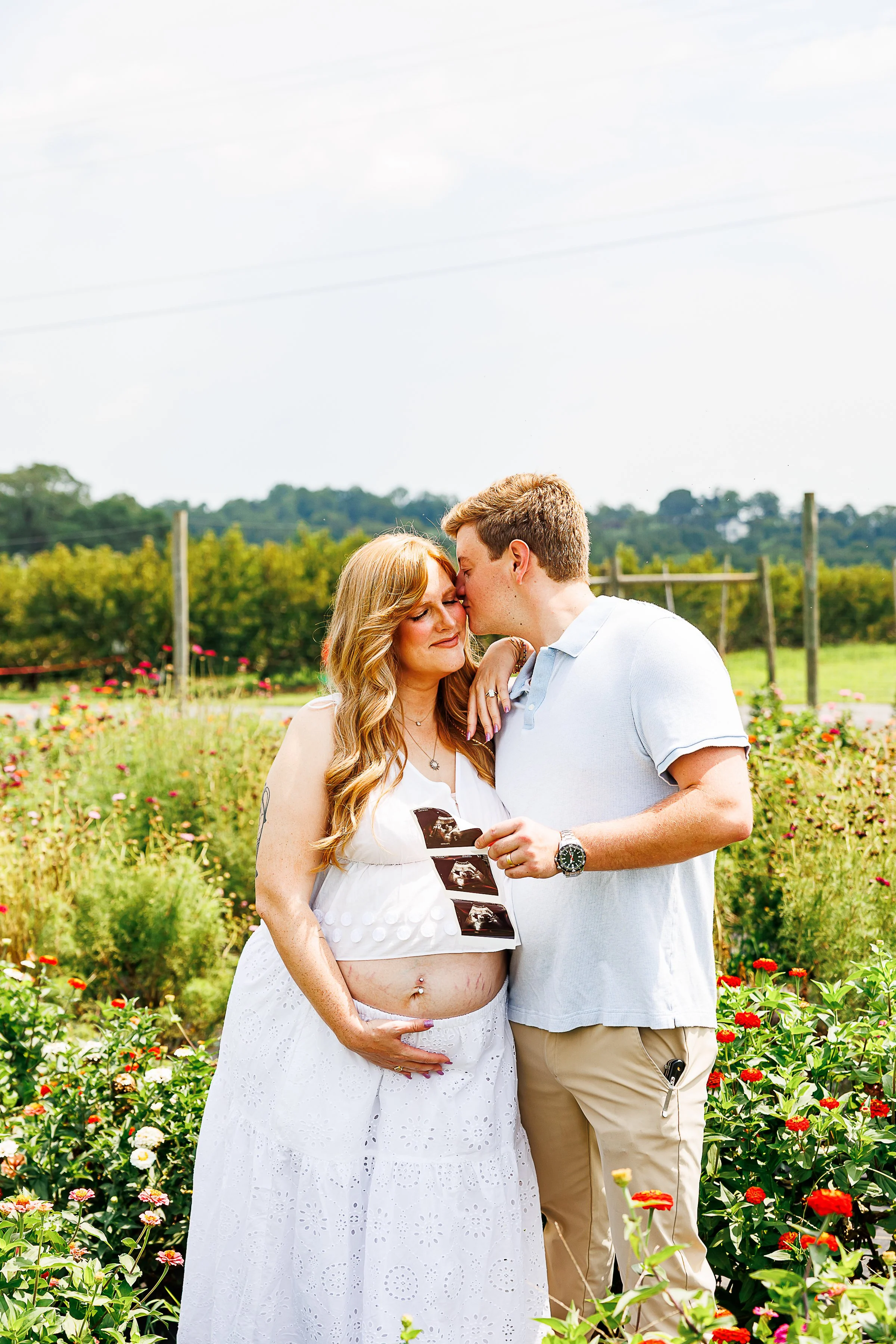 Couple standing together in a flower field with orchard rows in the background, holding ultrasound photos to the belly, partner kissing her forehead, at Chiles Peach Orchard in Crozet, VA.