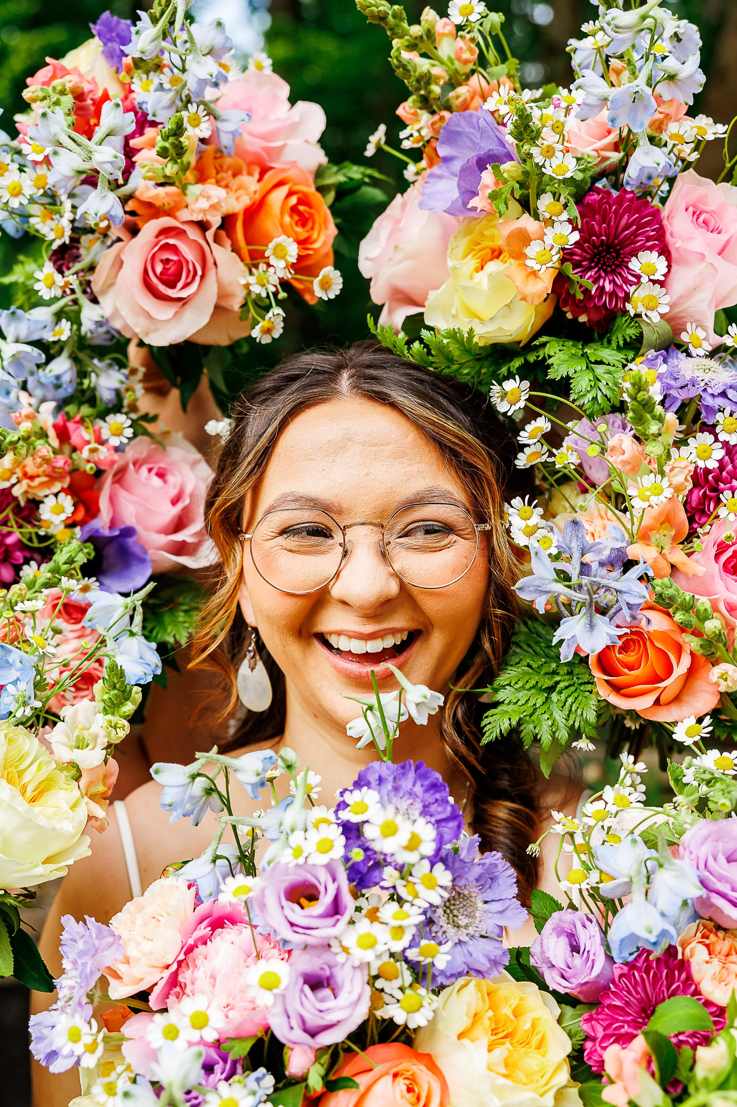 close-up portrait of a smiling woman with round glasses surrounded by vibrant, colorful floral arrangements. bright roses, daisies, delphiniums, and pastel blooms frame her face in shades of pink, orange, purple, yellow, and white. the joyful express