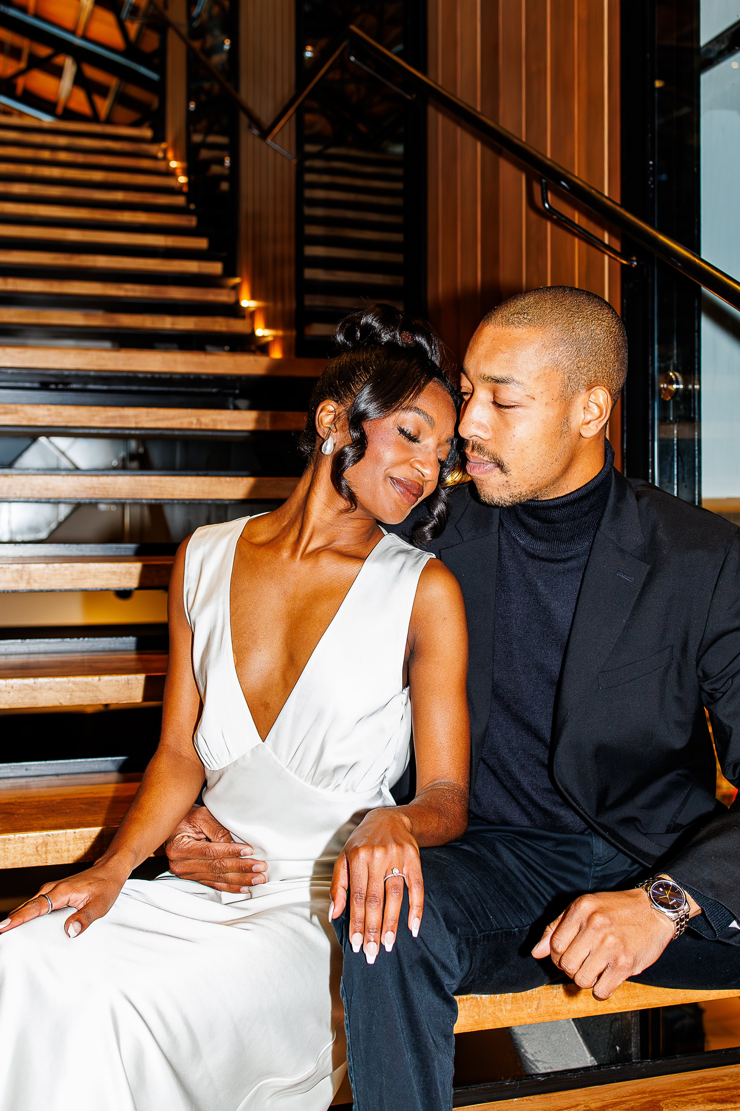 Couple sharing a quiet, close moment on a wooden staircase, her in a white satin dress resting her head near his shoulder, both dressed up for an evening out at Main Street Station in Richmond, VA.