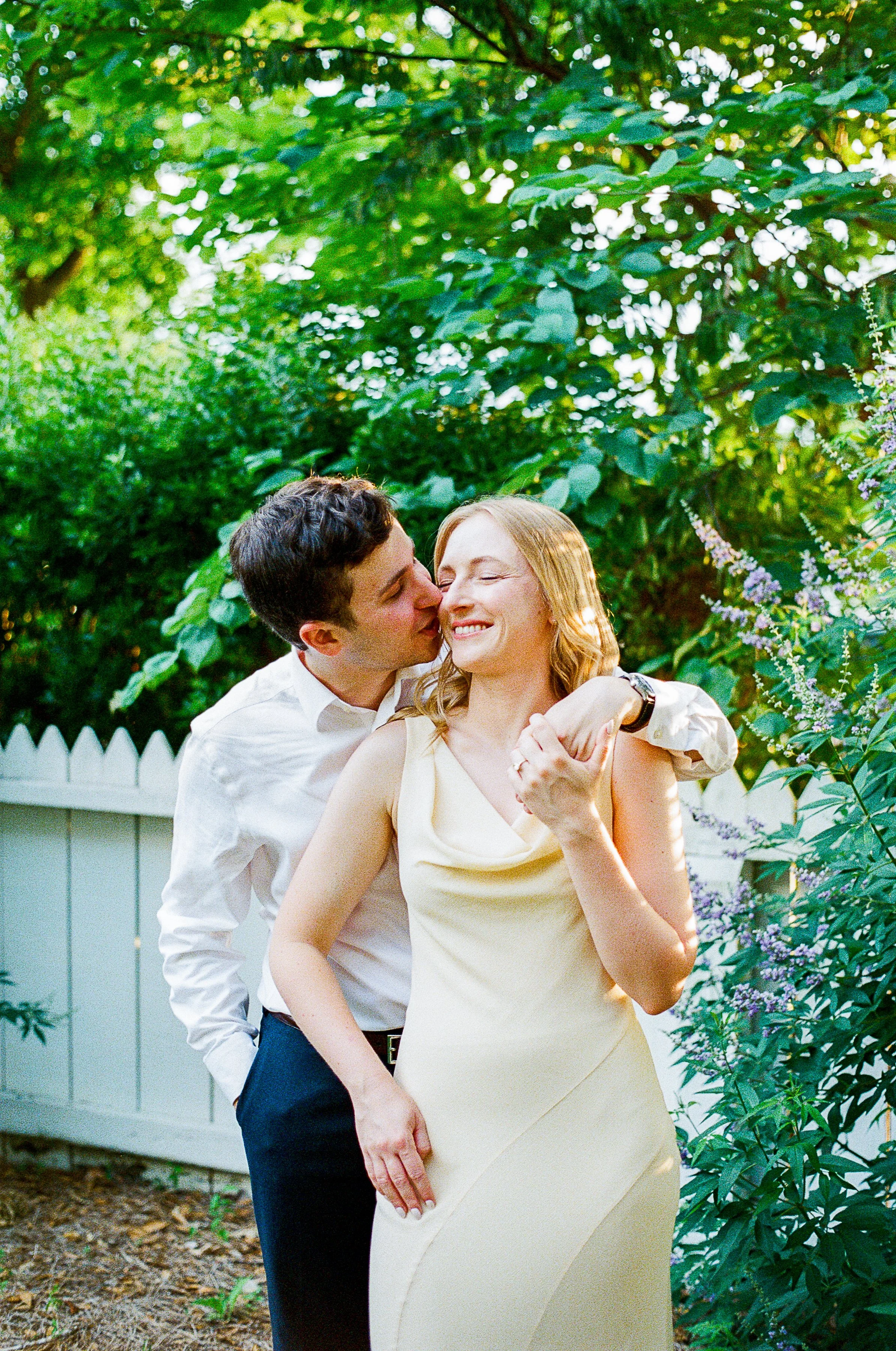 A couple shares a quiet, affectionate moment in a lush garden. One partner stands behind the other with an arm wrapped around their shoulders while leaning in for a kiss on the cheek. The other smiles with eyes closed, holding their partner’s hand. A