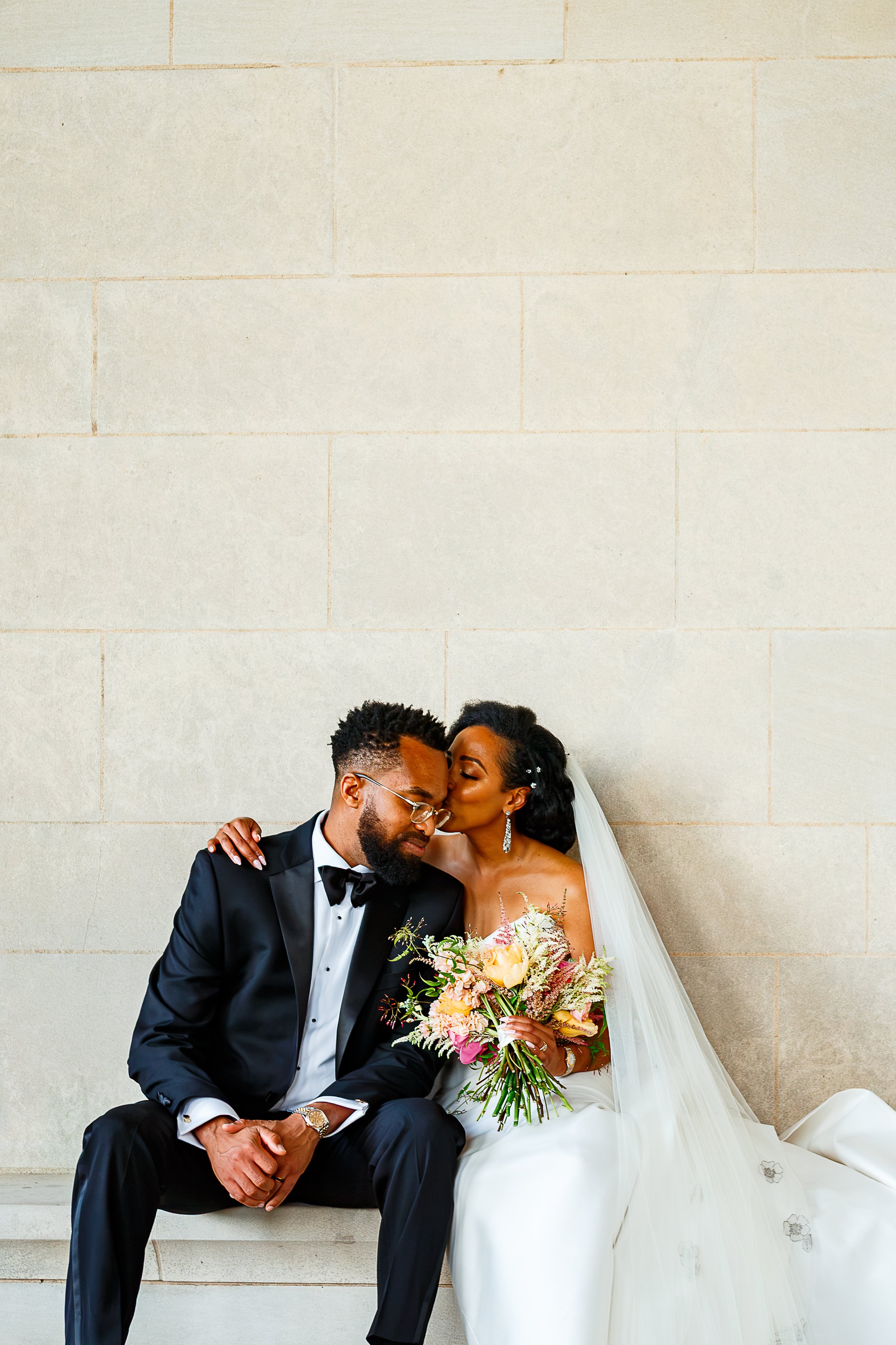 Bride in a strapless white gown carrying a bouquet of yellow roses and pink wildflowers, smiling up at the groom in a black tuxedo, walking a garden path beside the Gothic stonework of the National Cathedral in Washington, DC.
