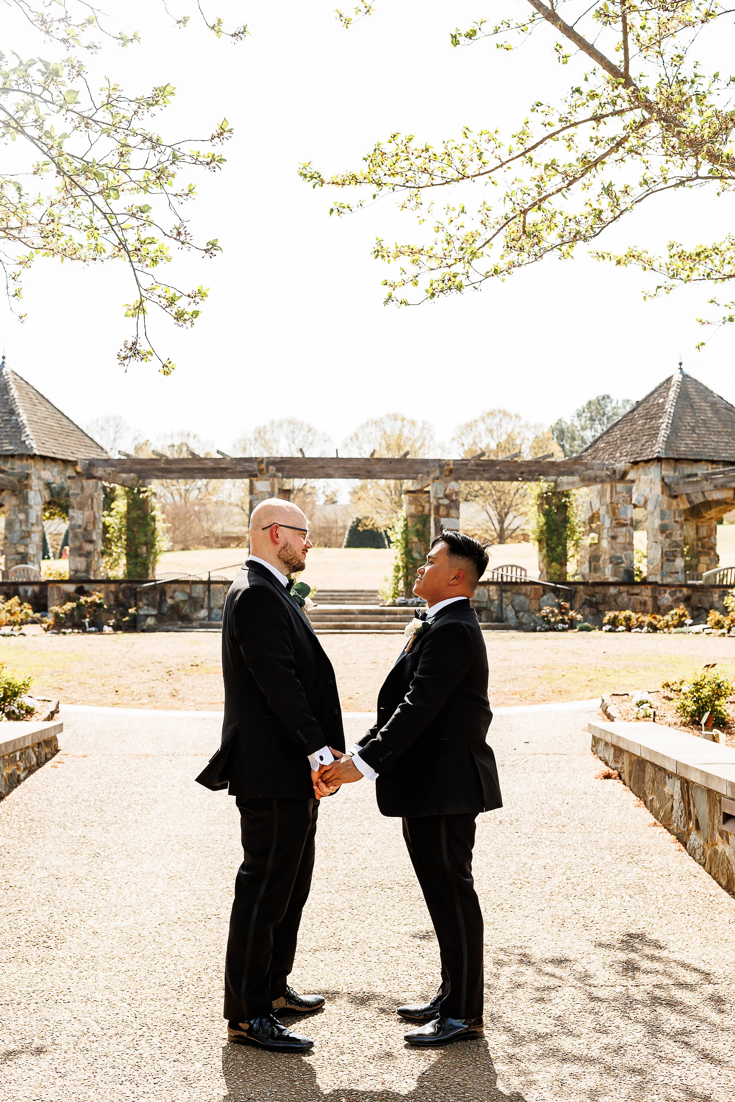 Two grooms in black tuxedos stand facing each other and holding hands in an outdoor garden setting, framed by stone archways and soft spring light filtering through tree branches above.