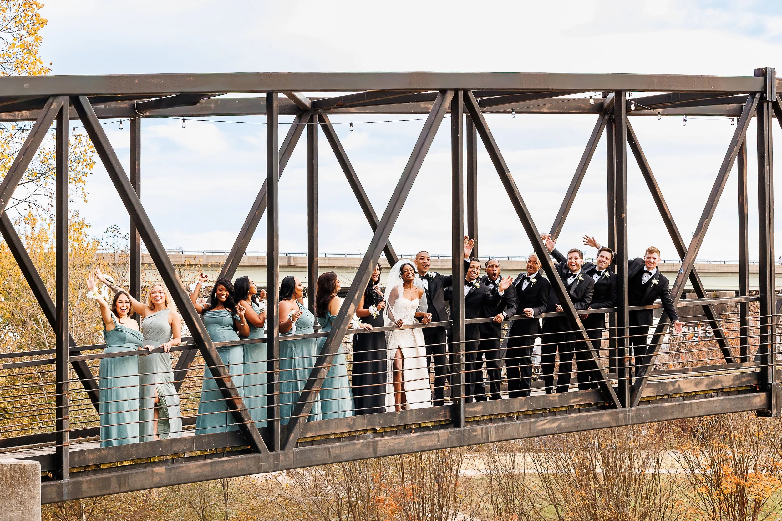 A joyful wedding party stands across a modern metal pedestrian bridge, with bridesmaids in sage green dresses on the left and groomsmen in black suits on the right. The bride, dressed in white, stands at the center smiling as the group cheers and wav