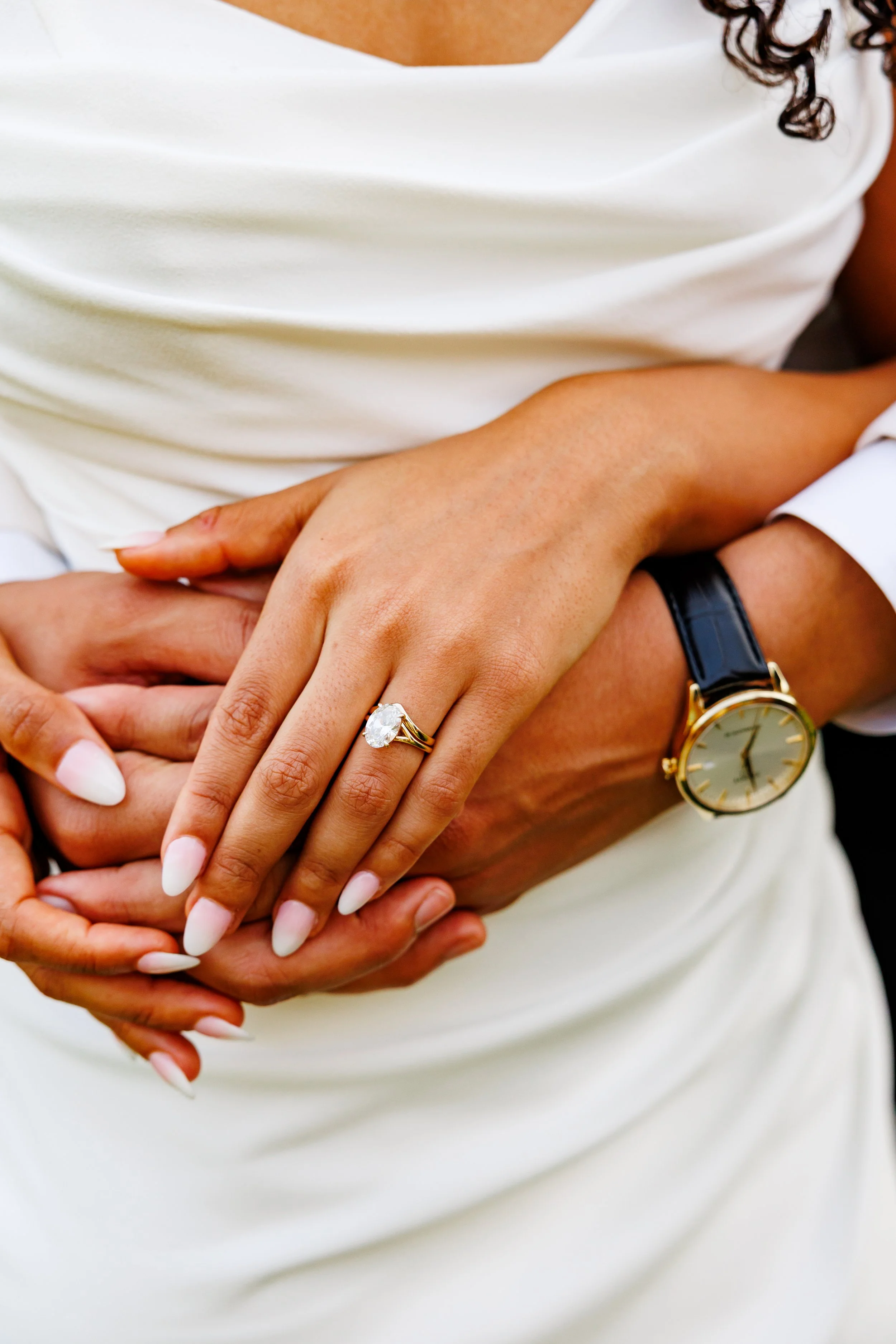 Close-up of a person wearing a gold watch and a diamond engagement ring, holding hands with another person against a white fabric background.