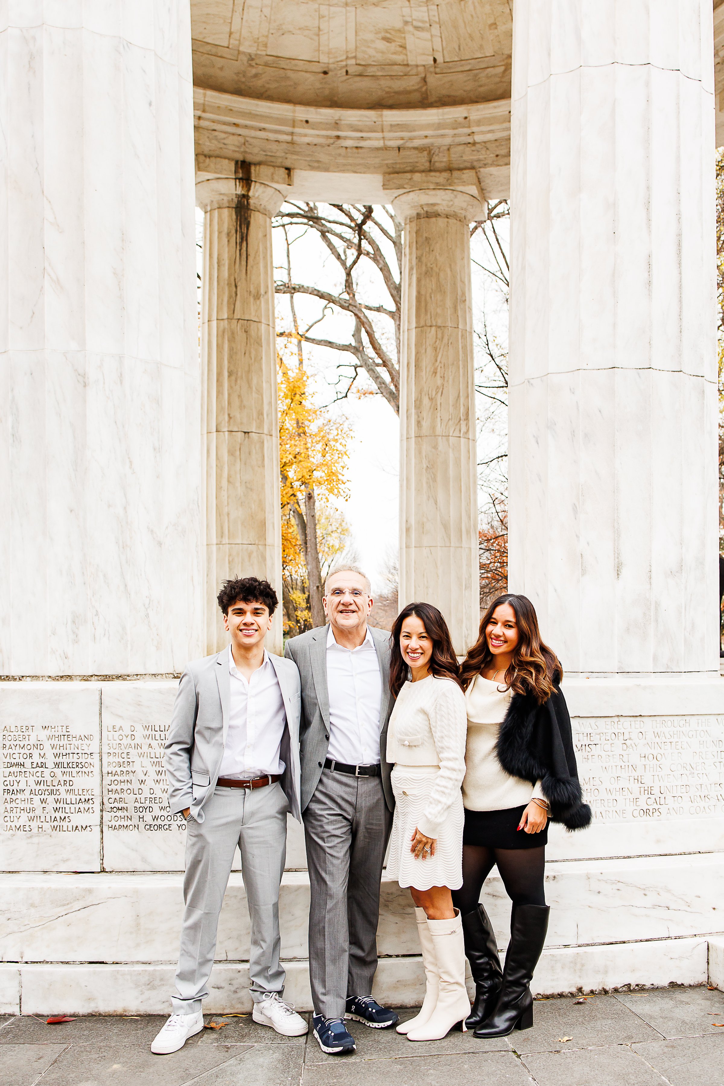 Family of four dressed in coordinated neutral tones - gray suits, cream sweaters, black boots — smiling together beneath the marble columns of the DC War Memorial in Washington, DC.