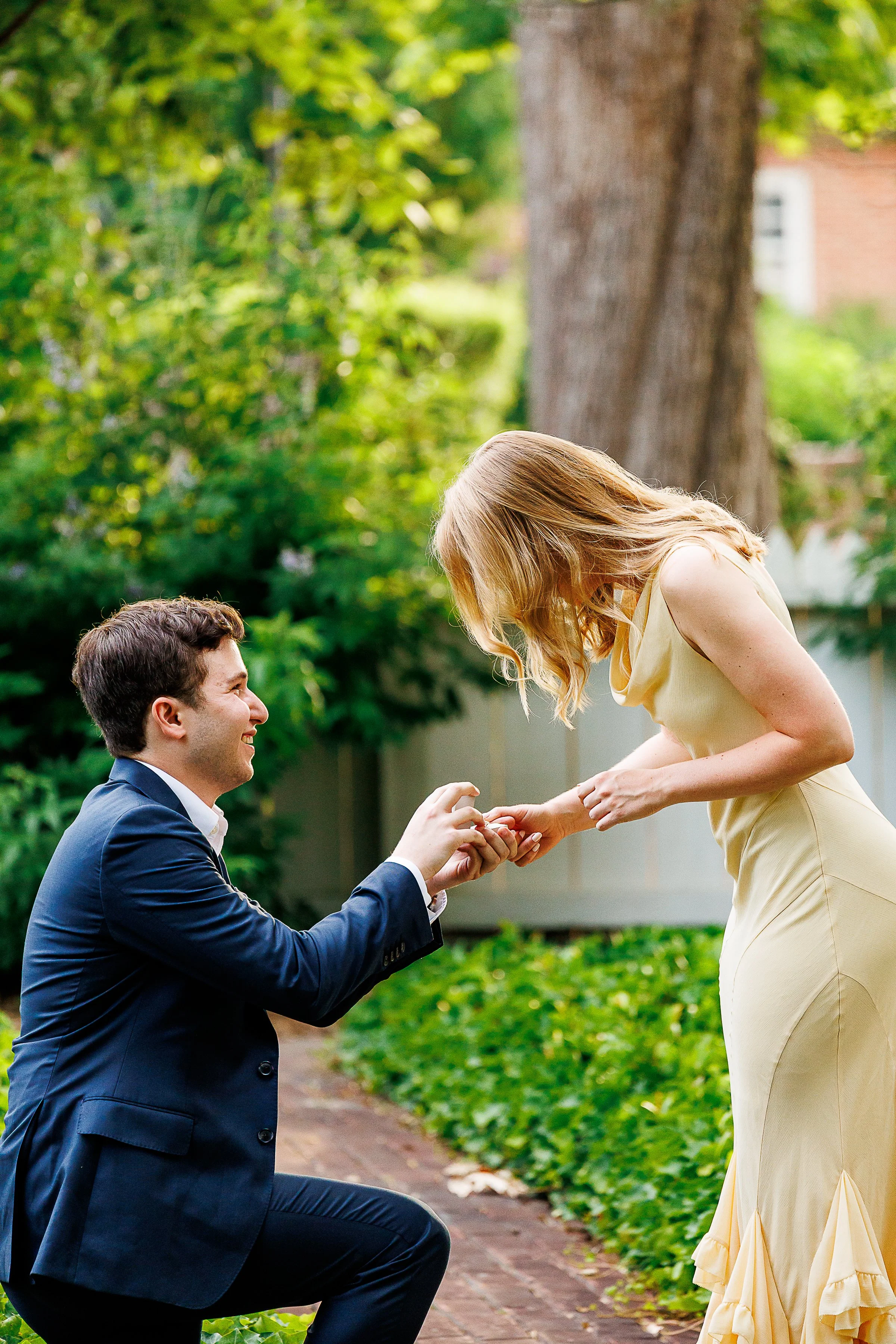A man in a blue suit kneeling and proposing to a woman in a light yellow dress outdoors with trees and greenery in the background.
