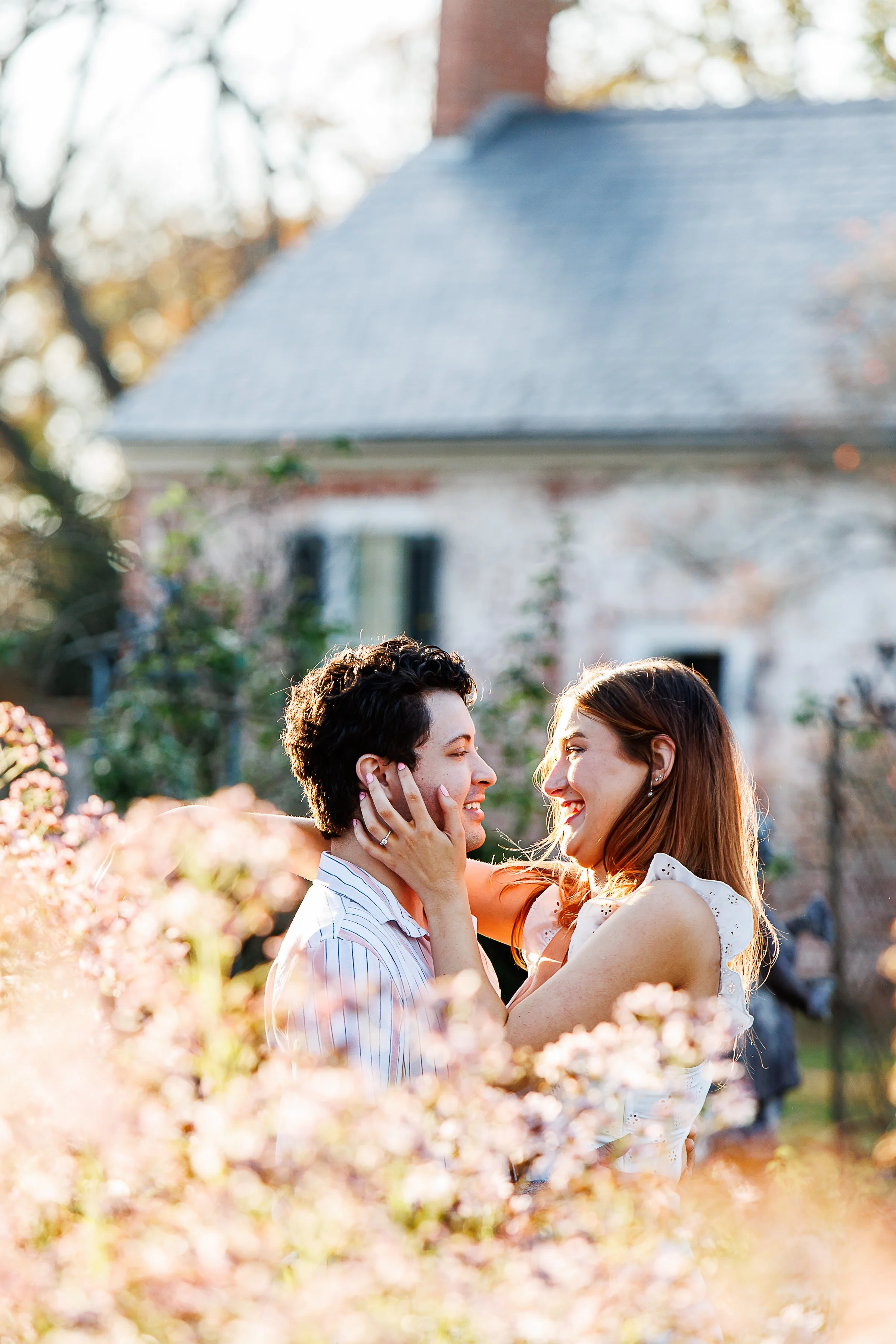 Couple smiling and embracing among blush cherry blossoms, her hand on his cheek, with a historic brick-and-stucco manor house in the background at Chatham Manor in Fredericksburg, VA.