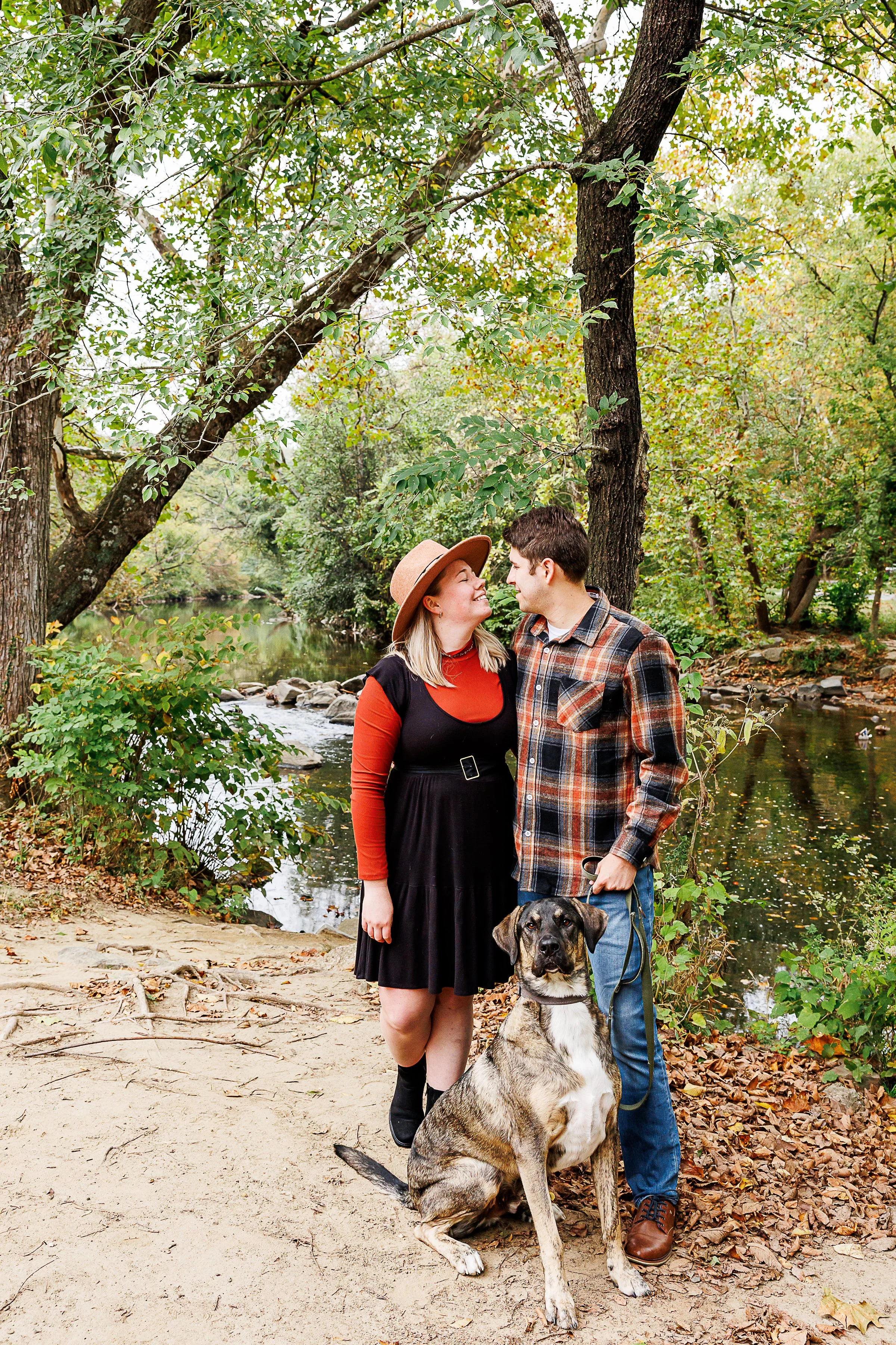 Couple standing close together with their dog beside a wooded creek, fall leaves scattered along the bank, at Rock Creek Park in Washington, DC.