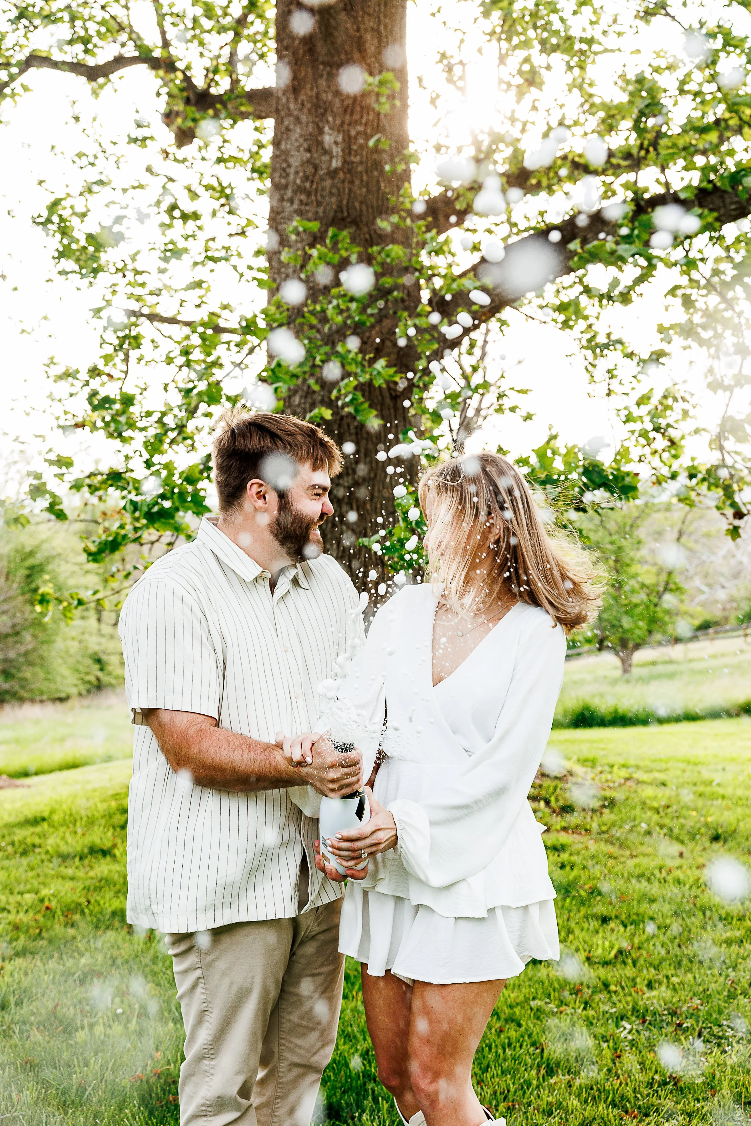 Couple laughing together as champagne sprays between them, her in a white ruffled dress and him in a striped shirt, under a large shade tree in a sunlit field in Loudoun County, VA.