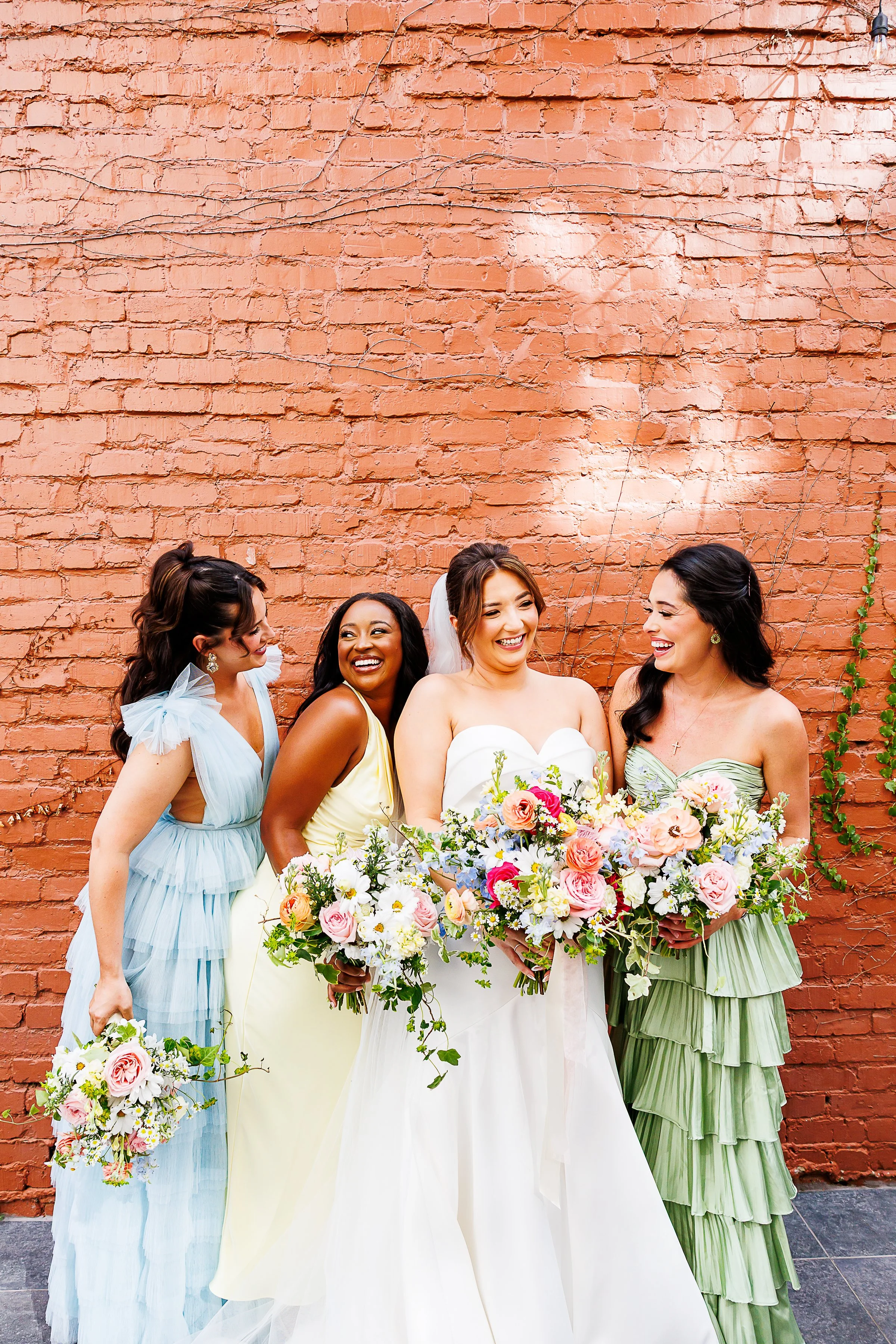 Bride and three bridesmaids in pastel blue, yellow, and sage green dresses laughing together with colorful bouquets against a brick wall at Common House in Richmond, VA.