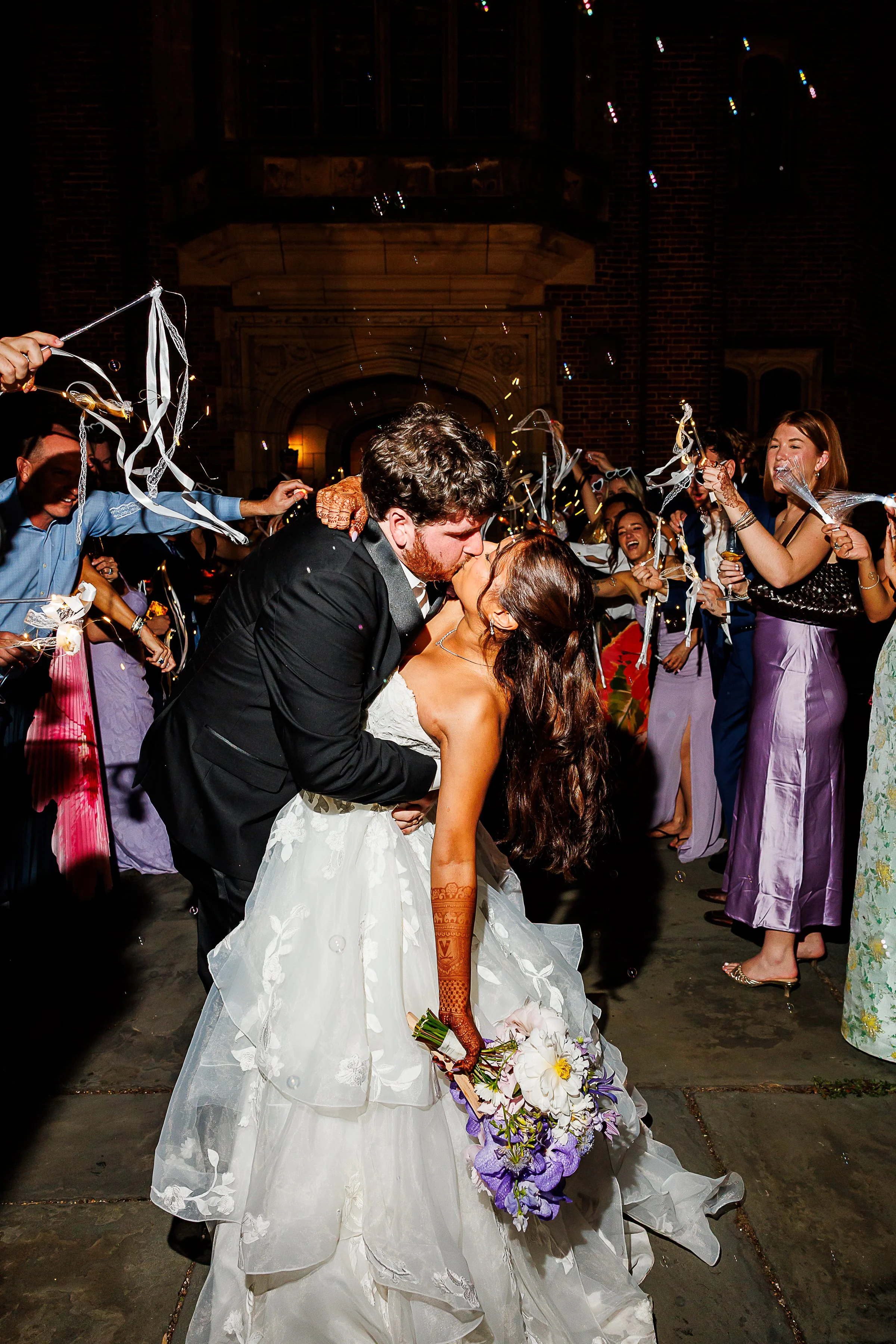 A newlywed couple shares a dramatic kiss outside a brick venue as friends surround them with ribbon wands and bubbles. The bride holds a colorful bouquet and wears intricate henna on her arms, while the groom wears a black suit.