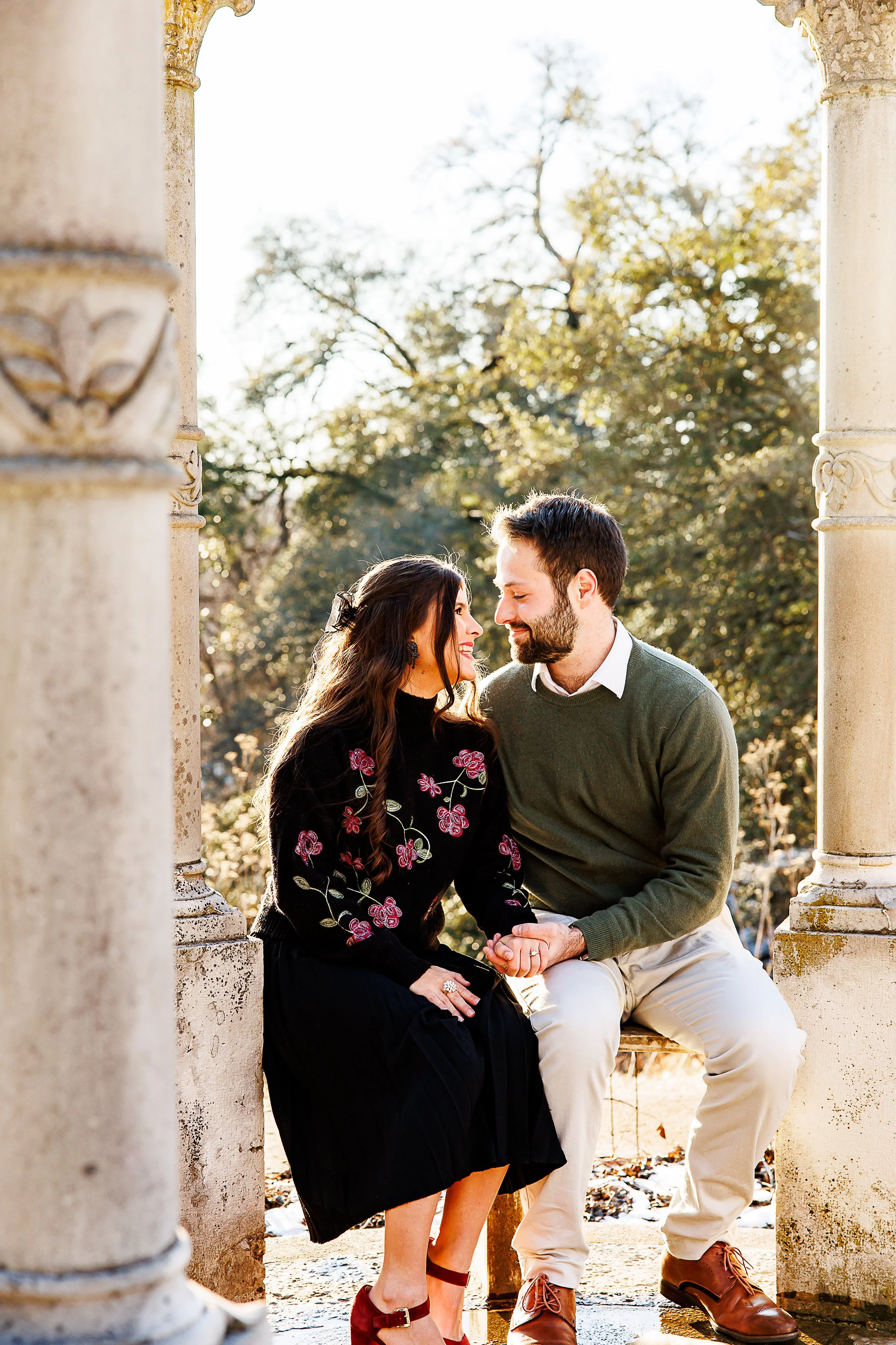 A couple sitting and facing each other between two stone columns outdoors, holding hands and smiling at each other with trees in the background.