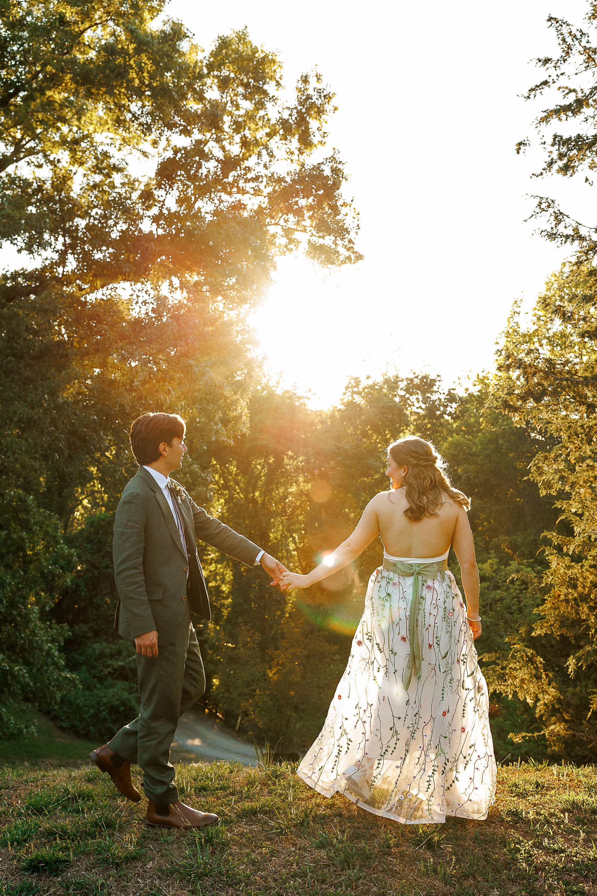 golden hour portrait of a couple holding hands on a grassy hill surrounded by tall trees and glowing sunset light. the groom wears a green suit with brown boots, while the bride wears a flowing backless floral wedding dress with embroidered details a