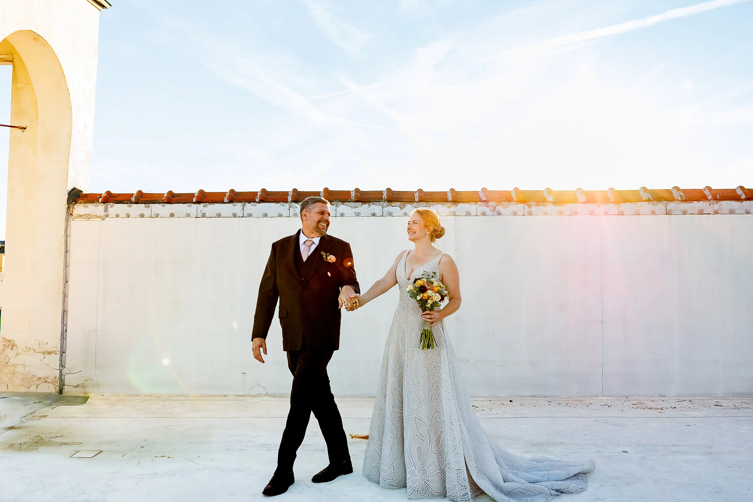 A newlywed couple walks hand in hand across a sunlit rooftop, smiling at each other as golden light spills over them. The bride holds a vibrant bouquet, her gown flowing behind her, while the groom in a dark suit matches her pace. A white stucco wall