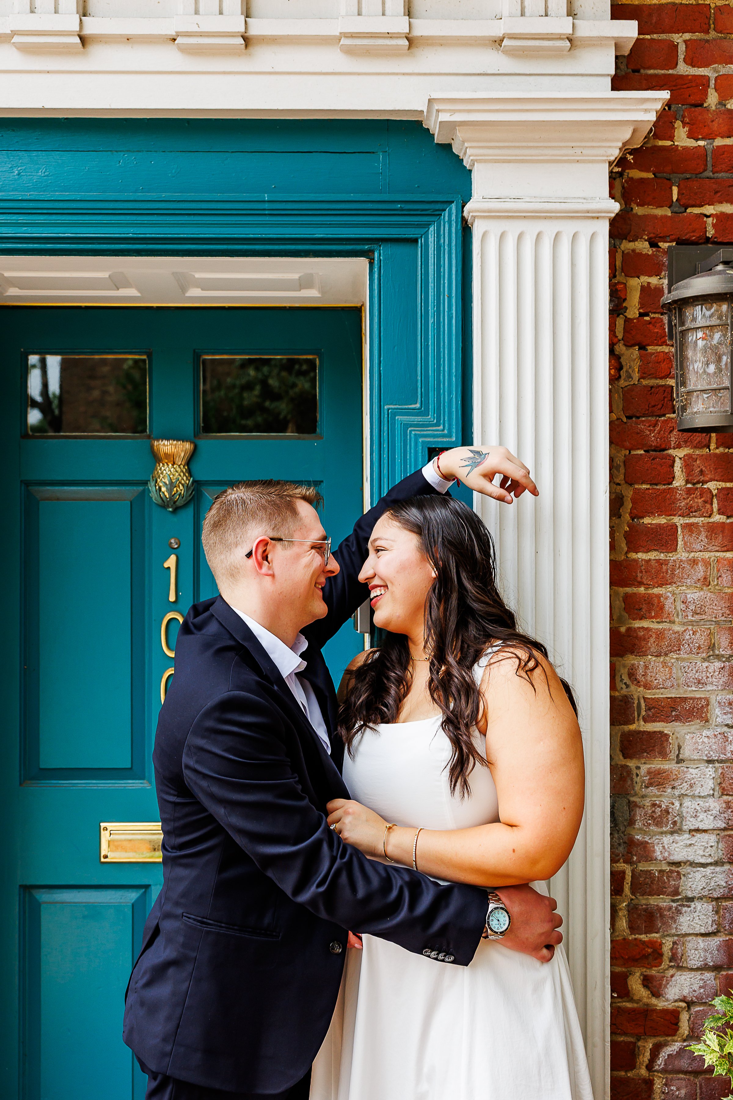 A couple shares a laugh outside a historic brick building with a bright teal front door. One partner leans against the doorway with an arm raised overhead while the other wraps an arm around their waist. They smile warmly at each other in a candid, j
