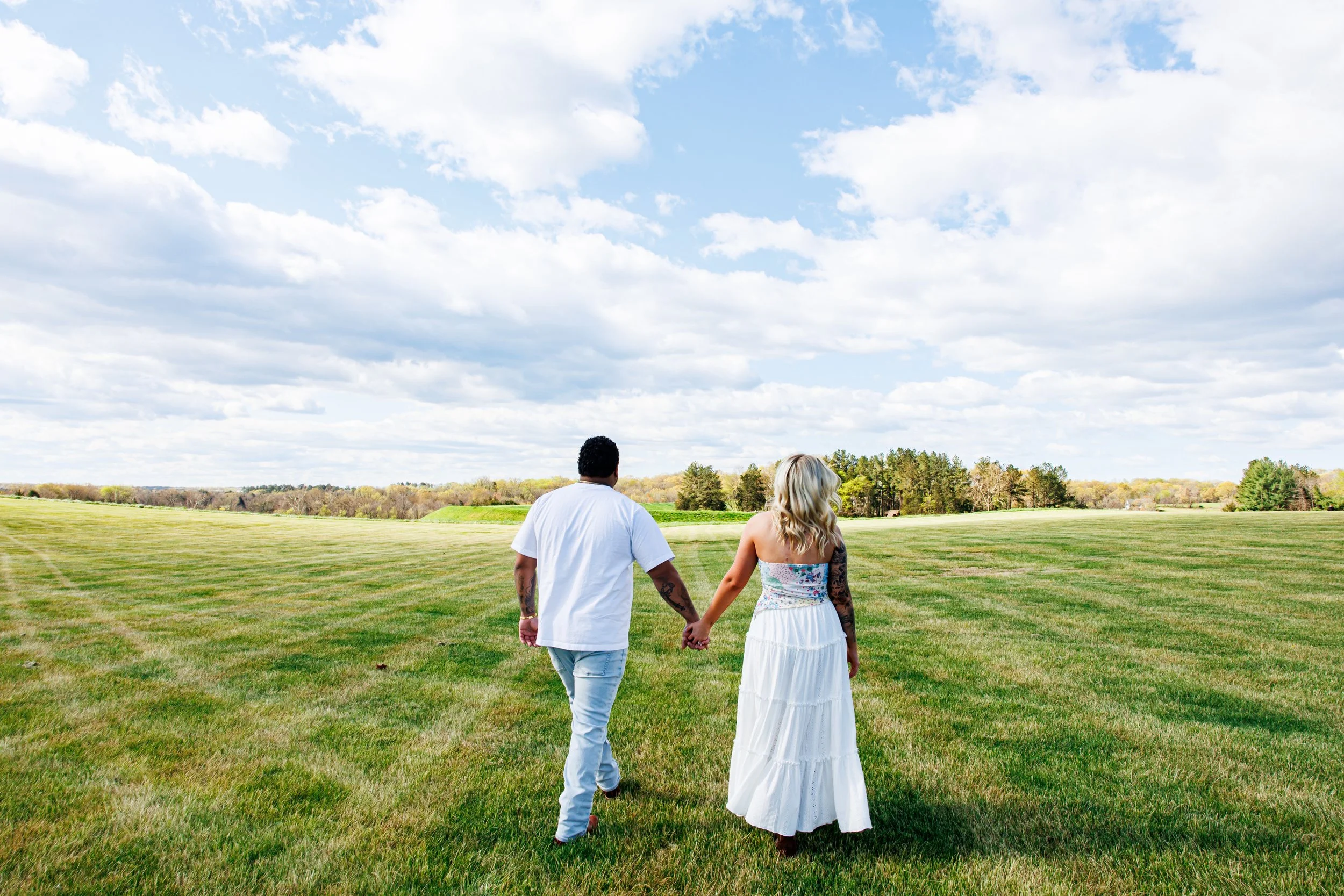 romantic outdoor portrait of a couple holding hands while walking through a wide open grassy field beneath a bright blue sky with fluffy clouds. the woman wears a white tiered dress with a floral corset top, and the man wears a white t-shirt and ligh