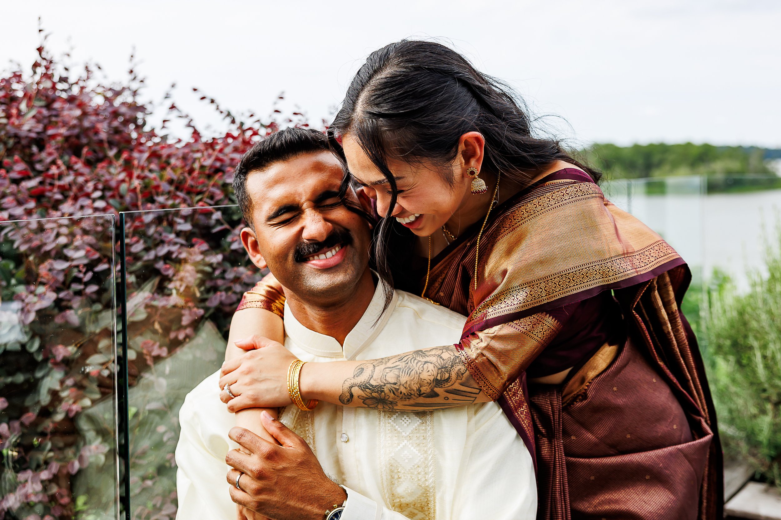 A couple shares a joyful embrace during a wedding celebration outdoors by the water. The woman, wearing a deep burgundy and gold silk sari with gold jewelry, wraps her arms around the man from behind as they both laugh with their eyes closed. The man