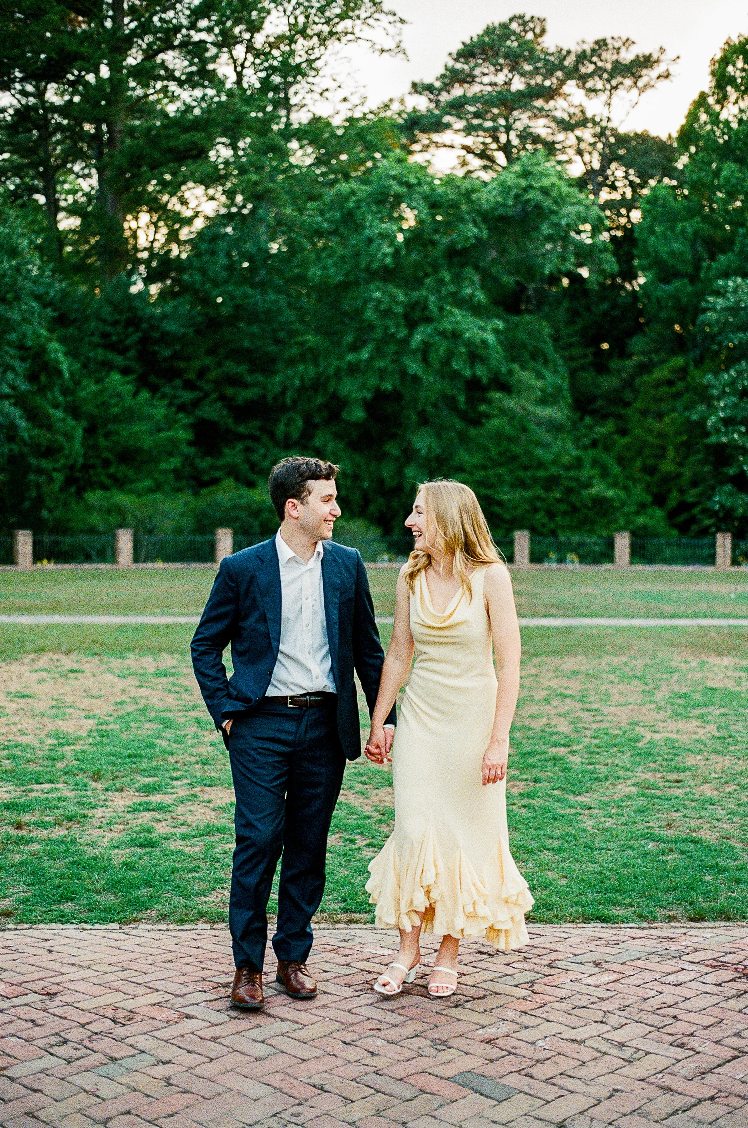 A couple walks hand in hand across a brick pathway, smiling warmly at one another. One wears a navy suit with a white dress shirt, while the other wears a flowing cream-colored midi dress and white heels. An open lawn and towering trees create a peac