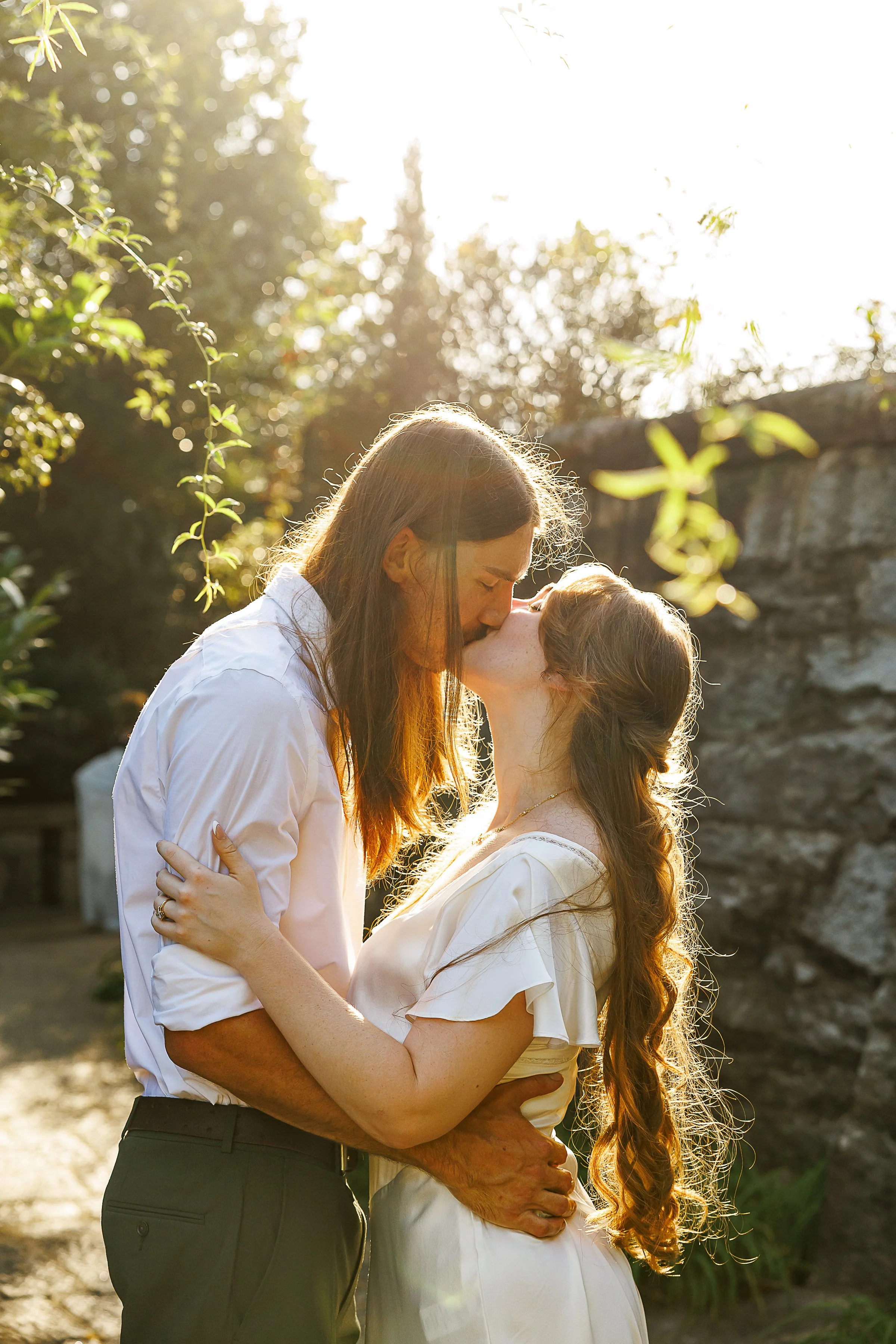 Bride and groom sharing a kiss in golden-hour sunlight in the Italian Garden at Maymont in Richmond, VA, her long hair catching the light against the stone wall backdrop.