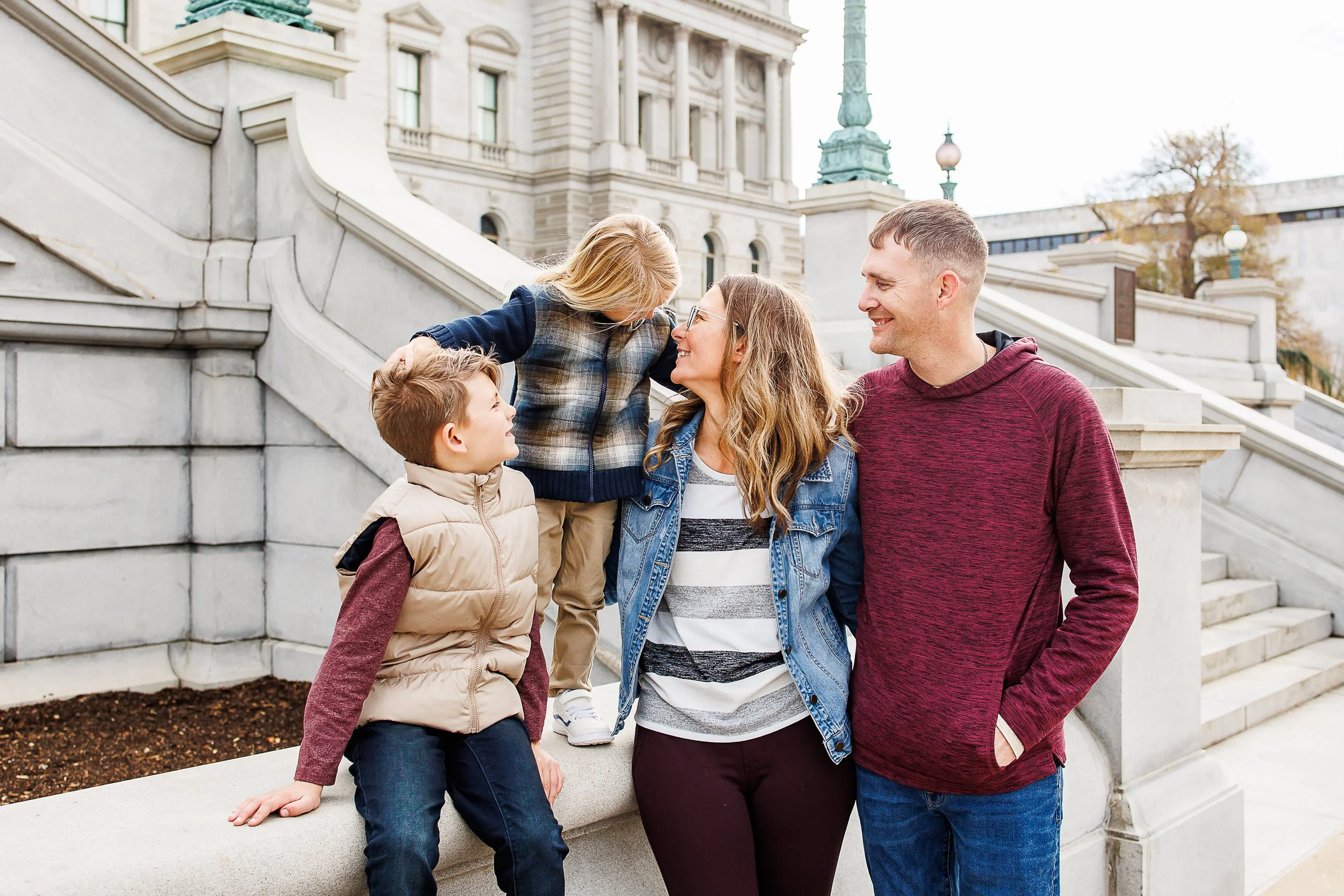 Family of four sharing a laugh on the marble steps of the Library of Congress, one son perched on the ledge and the other standing to be kissed on the forehead, on Capitol Hill, Washington, DC.