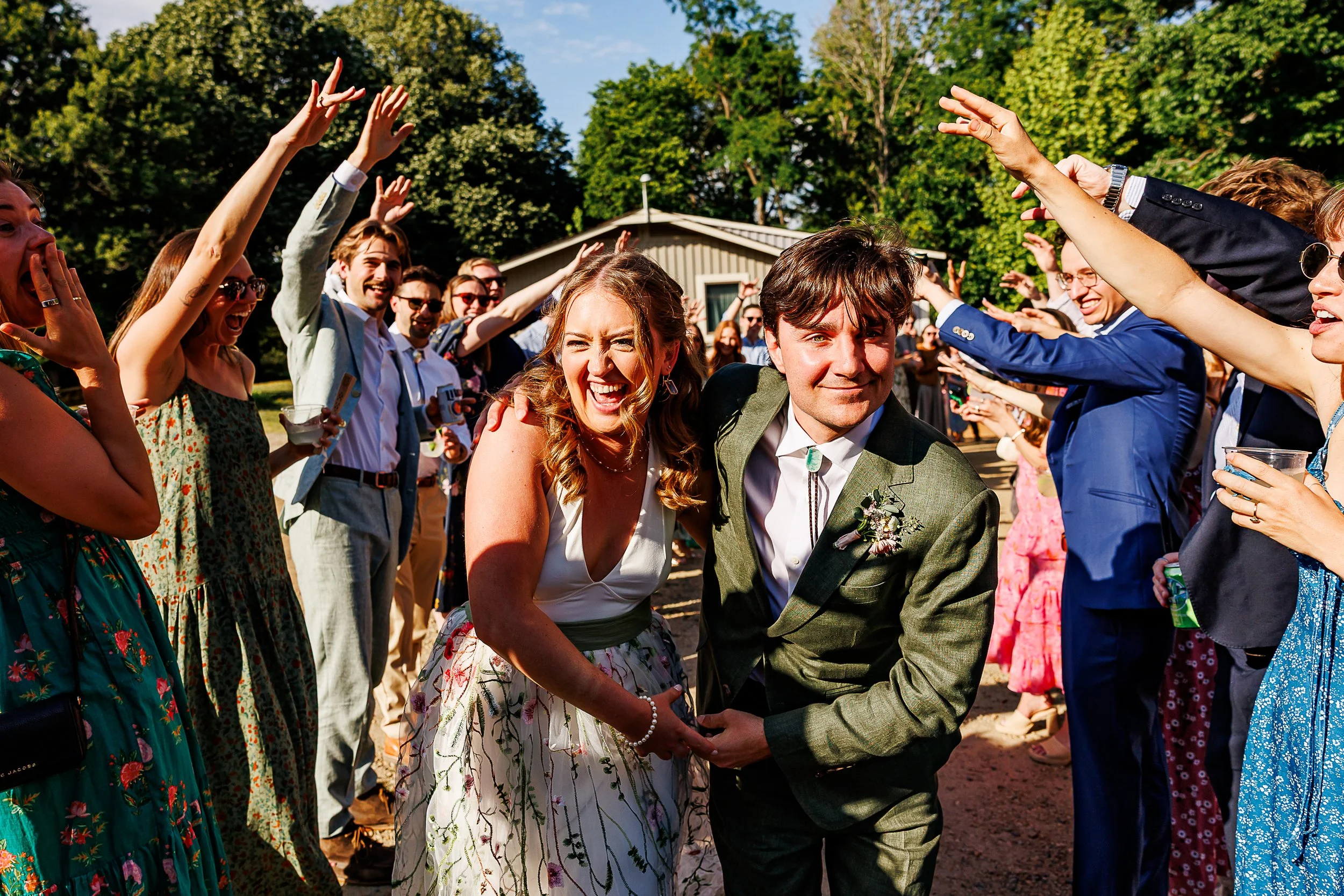 Newlyweds run hand in hand through a joyful tunnel of cheering wedding guests with their arms raised in celebration. The bride wears a sleeveless floral embroidered gown with a deep V-neckline, while the groom wears a textured olive green suit with a