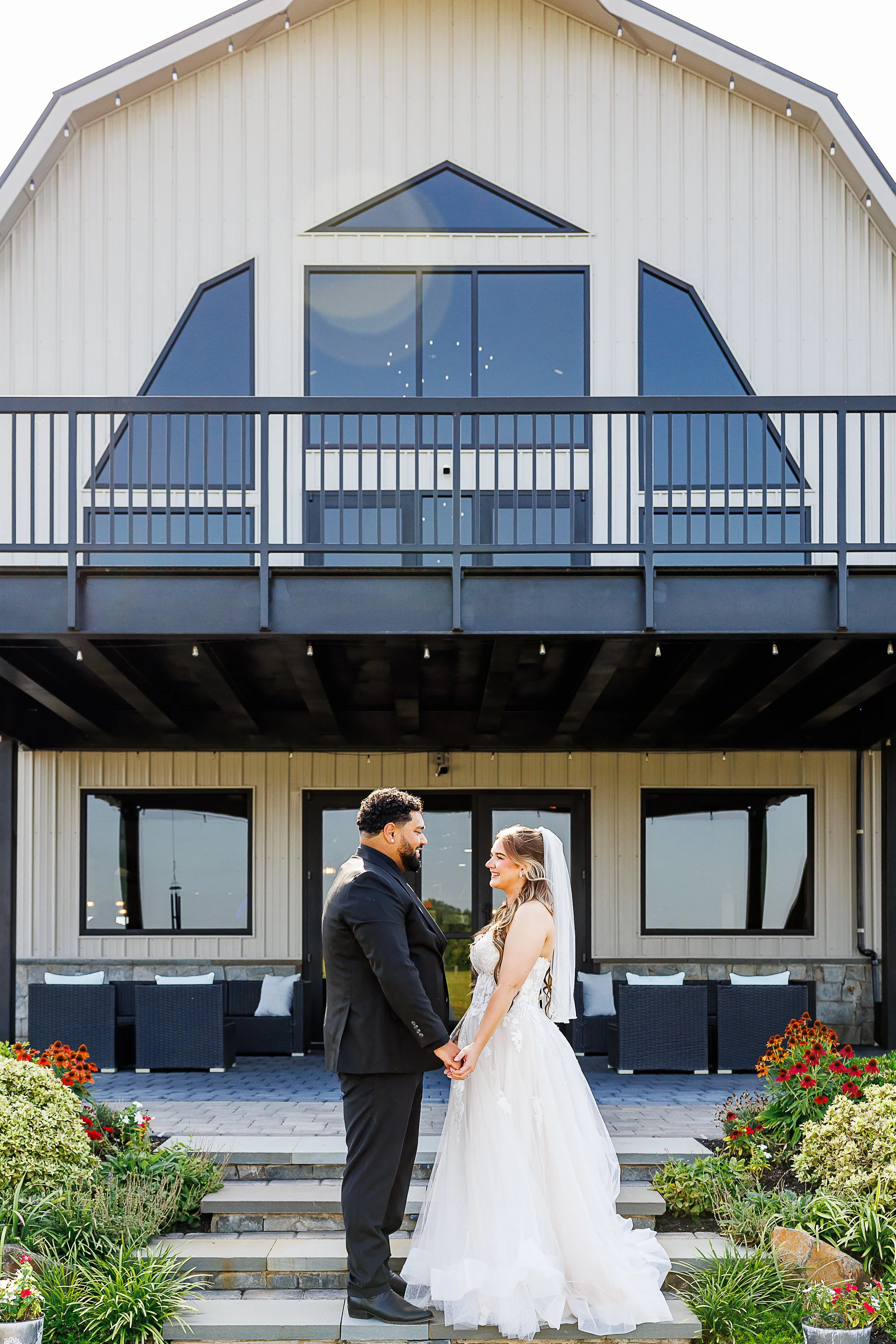Bride and groom stand hand in hand on the front steps of a modern white barn wedding venue, smiling at each other beneath the building's striking geometric windows and black balcony. Colorful flower beds frame the foreground while the clean architect