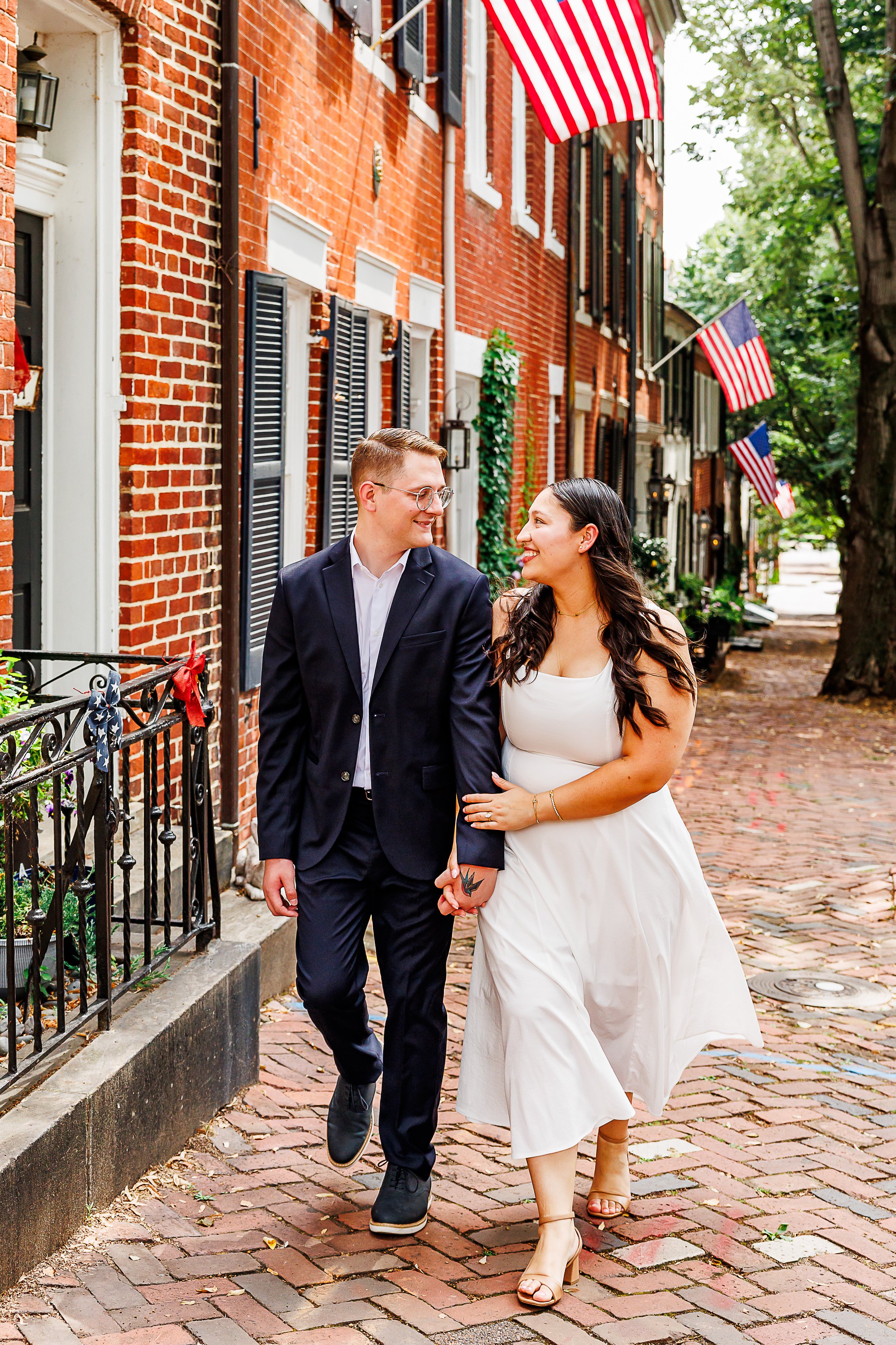 A couple strolls hand in hand down a brick-lined historic street, smiling at one another. One partner wears a navy suit while the other wears a flowing white dress and nude heels. American flags, red brick row houses, and leafy trees line the sidewal