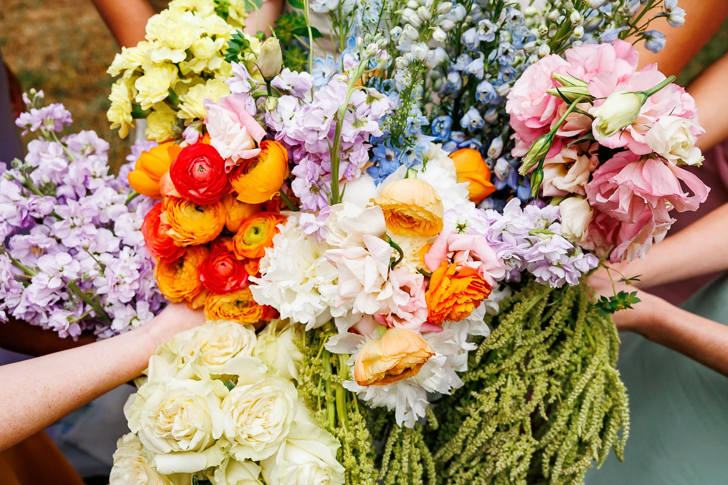 People holding a large colorful bouquet of mixed flowers, including ranunculus, lisianthus, delphinium, and stock, with green foliage in the background.