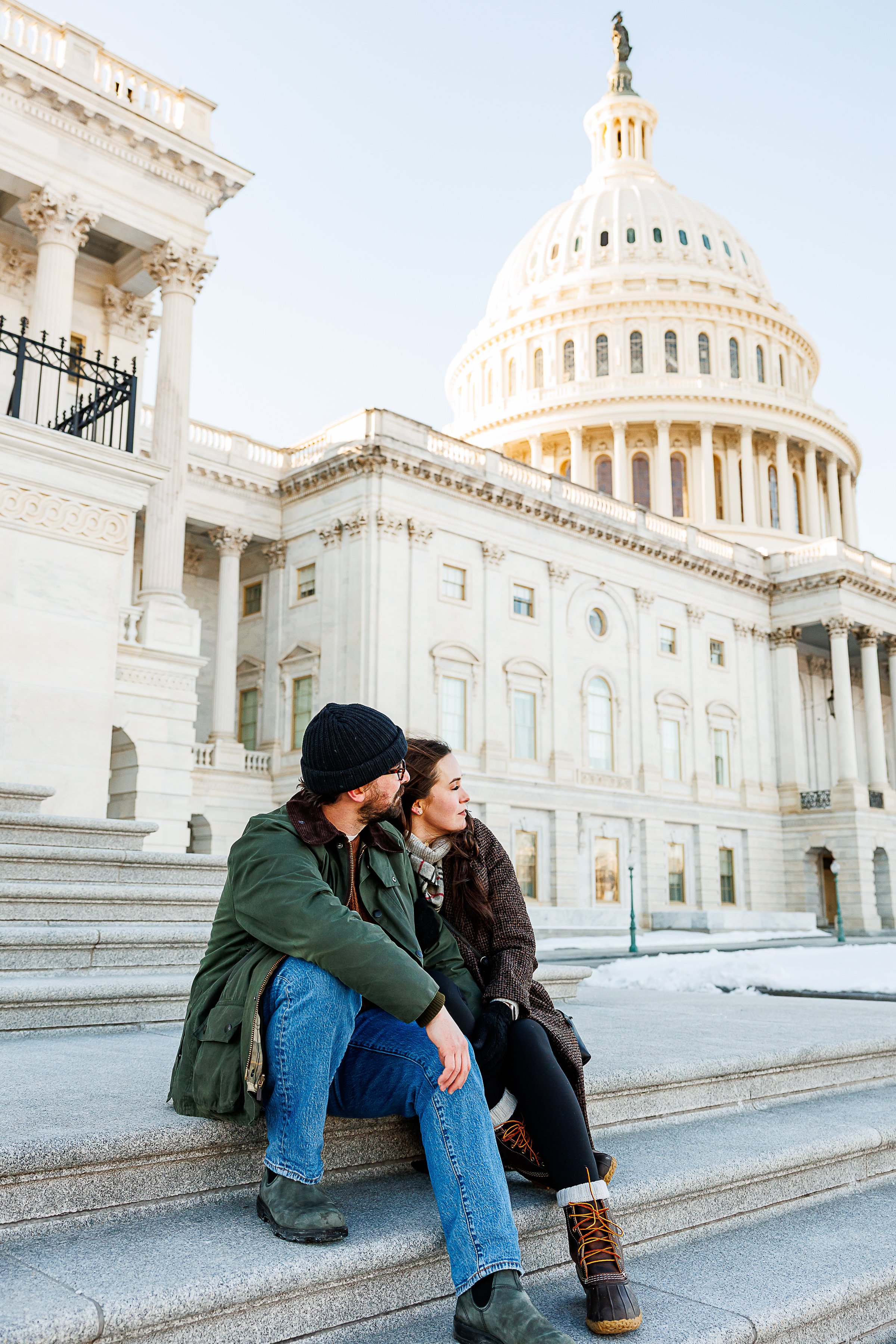 A couple sits closely together on the steps of the U.S. Capitol, bundled in winter layers as they take in the view. They lean into each other, sharing a quiet, affectionate moment against the grand backdrop of the Capitol’s white columns and dome, wi